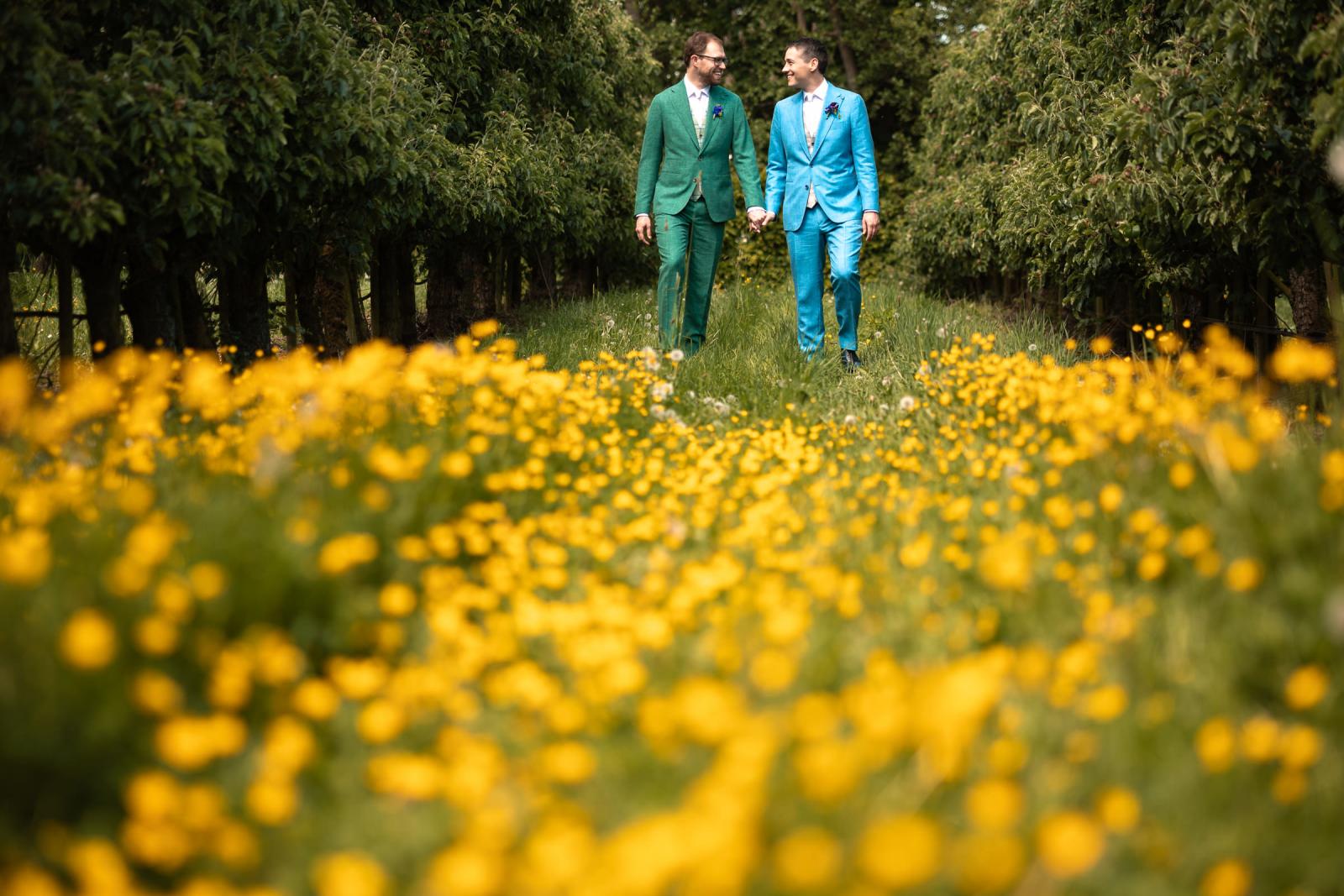 Daan en Martijn lopen glimlachend hand in hand over een graspad, gefotografeerd met een dromerige, onscherpe voorgrond vol gele boterbloemen