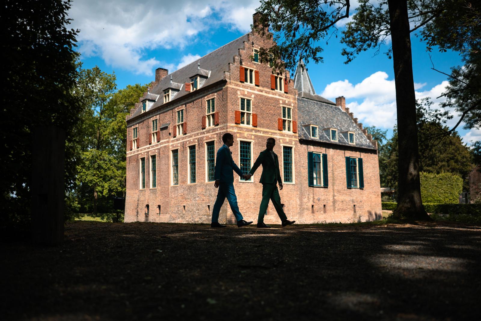 Bruidegoms Daan en Martijn lopen hand in hand over het terrein langs de monumentale, bakstenen gevel van Kasteel Wijenburg