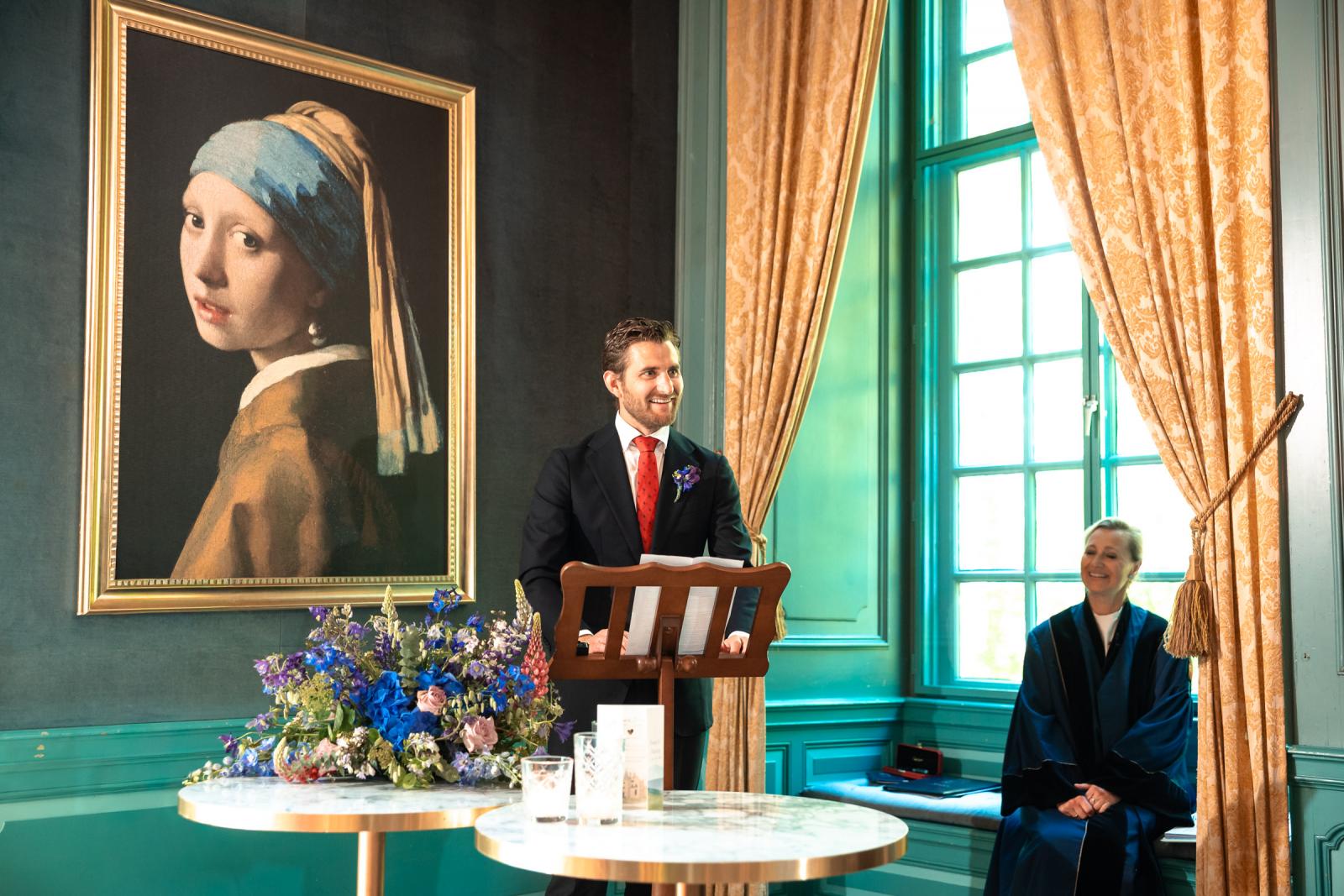 Een mannelijke daggast staat achter de katheder en houdt een speech naast een grote replica van het schilderij 'Het meisje met de parel