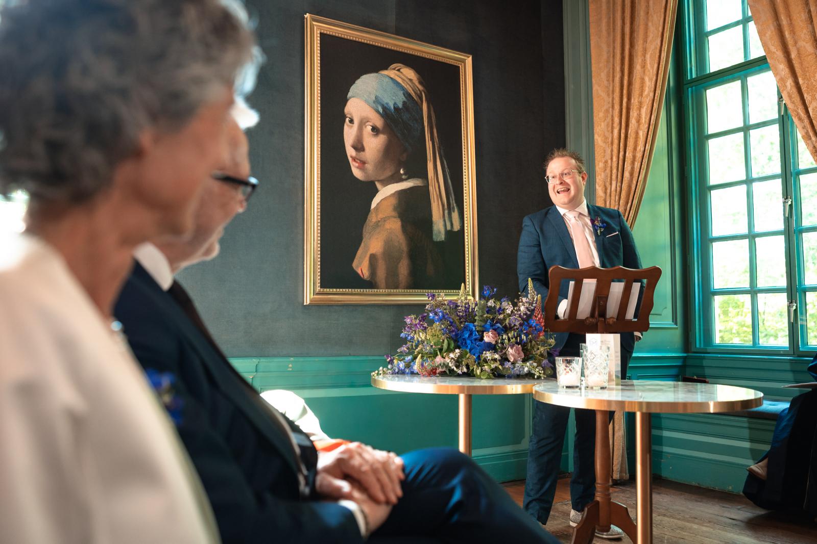 Een lachende gast in een donkerblauw pak spreekt het bruidspaar toe vanaf de katheder in de blauwe salon van Kasteel Wijenburg