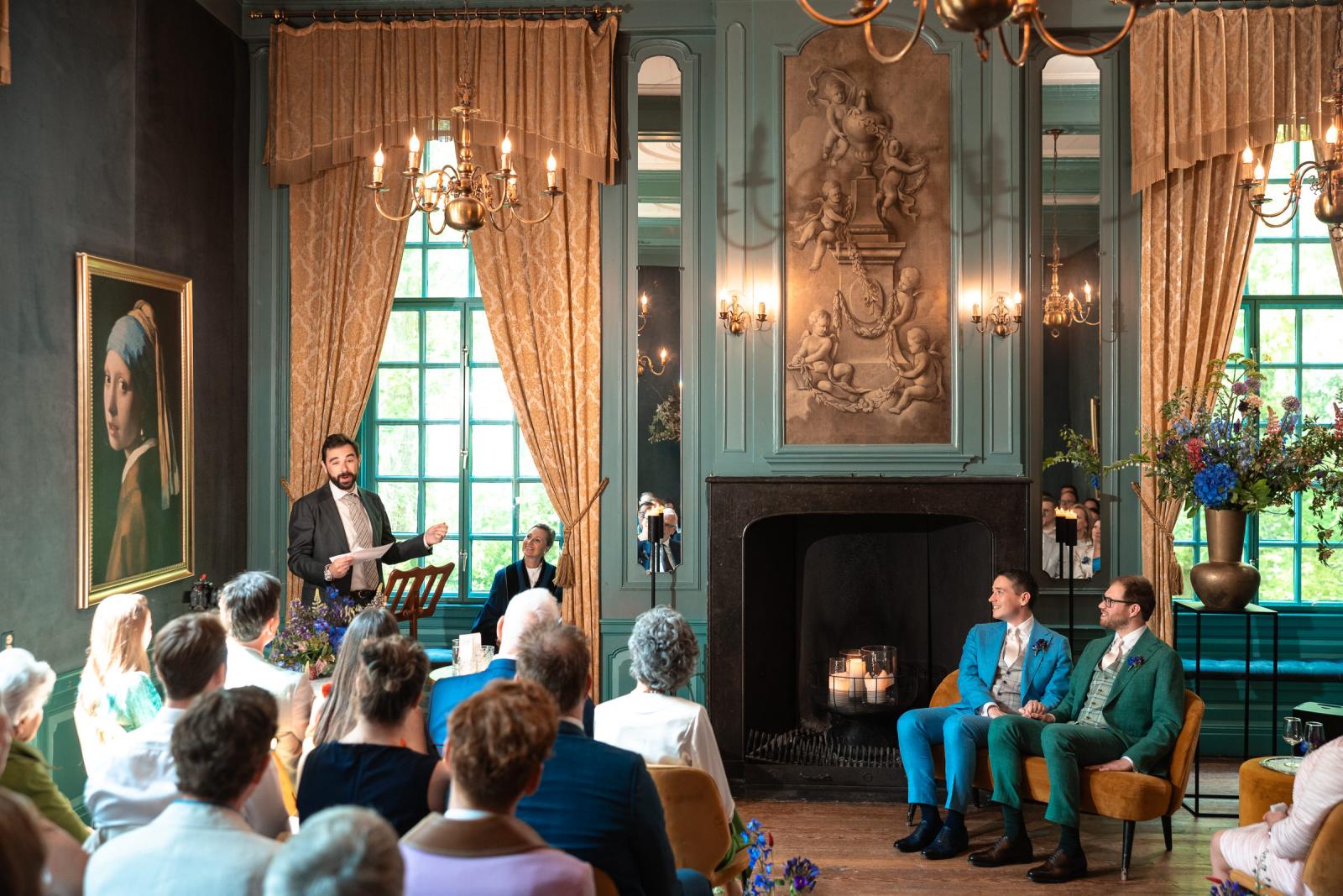 Daan en Martijn zitten hand in hand op een okergele fluwelen bank en luisteren glimlachend naar een speech van een van hun getuigen