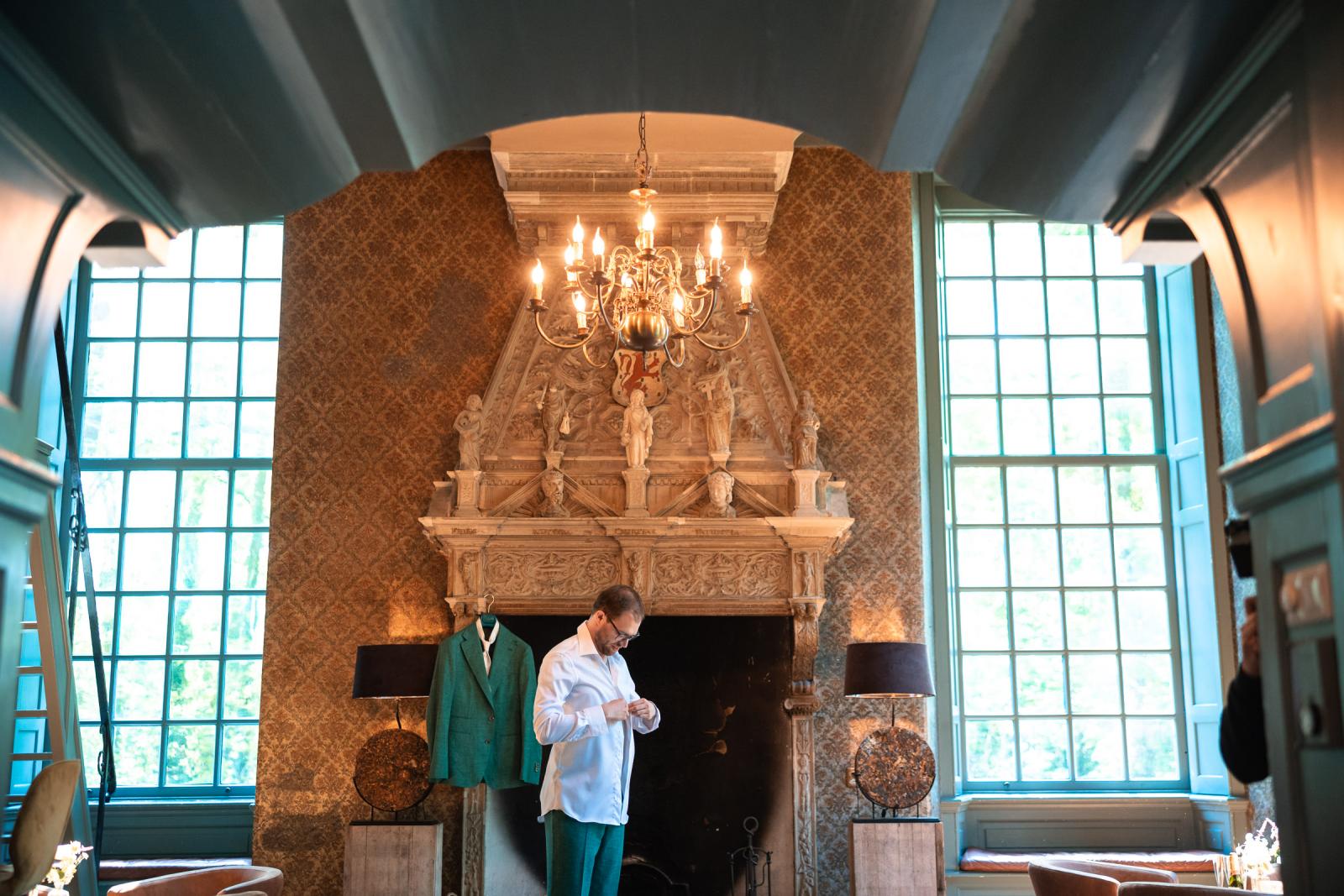 Bruidegom Martijn knoopt zijn witte overhemd dicht in een kasteelkamer met een rijk gedecoreerde schouw en een grote kroonluchter