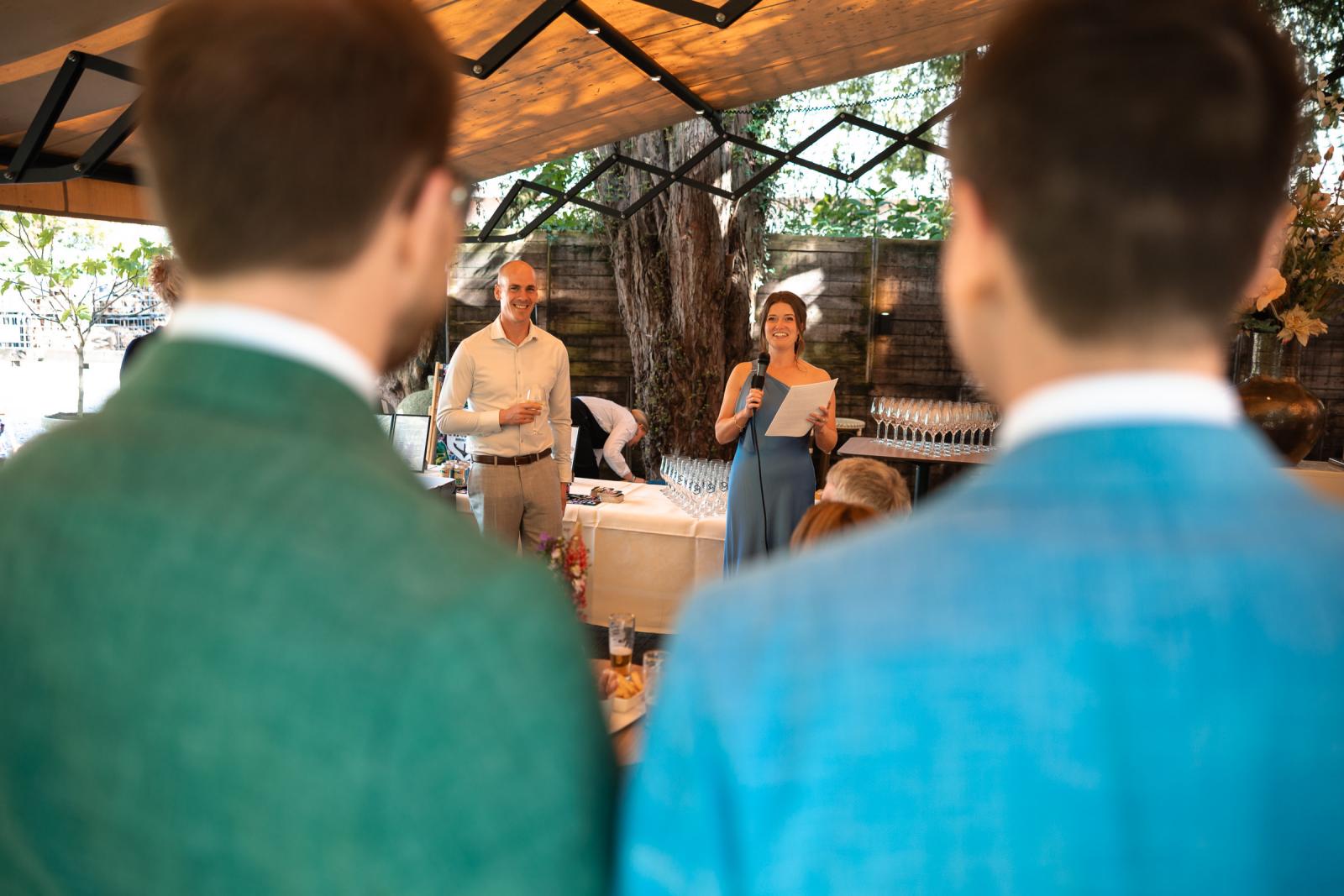 Zicht over de schouders van de bruidegoms naar een vrouwelijke gast in een blauwe jurk die een speech houdt door een microfoon