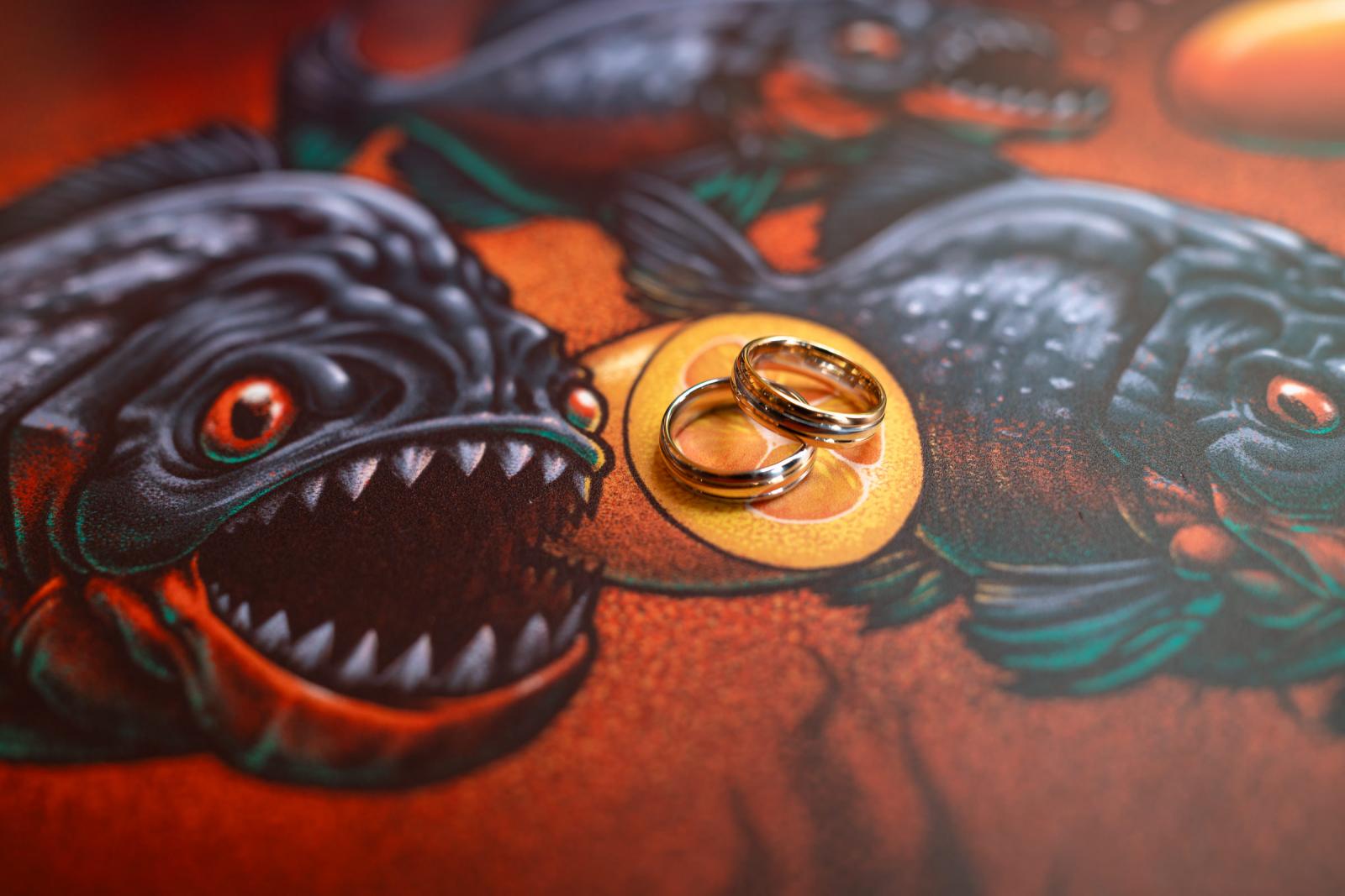 Close-up van de twee gouden trouwringen die op een kleurrijke ondergrond met een getekende piranha-afbeelding van een biermerk liggen