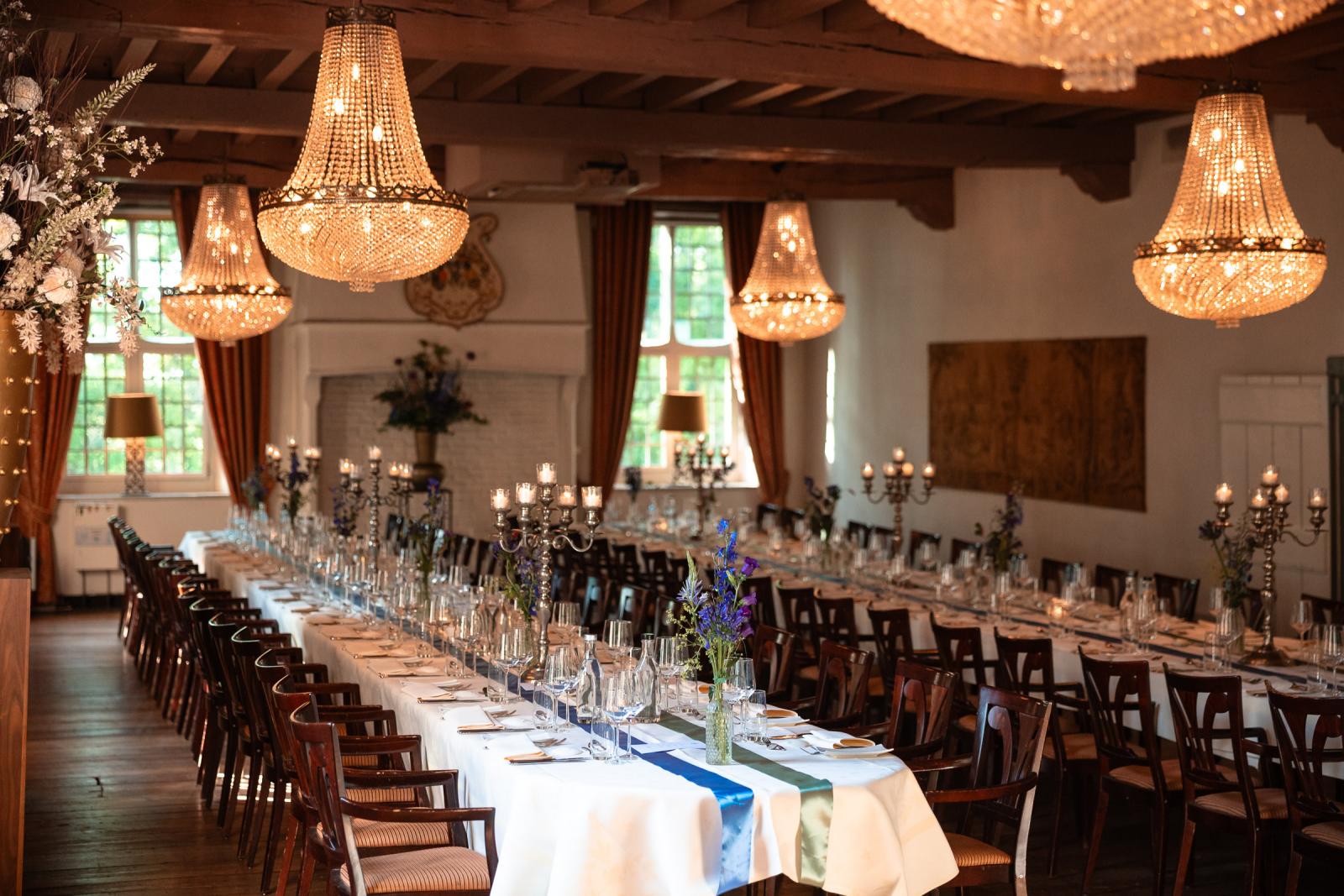 Overzicht van de sfeervolle dinerzaal met houten balkenplafond, verlicht door grote kristallen kroonluchters en kandelaars op de lange tafels