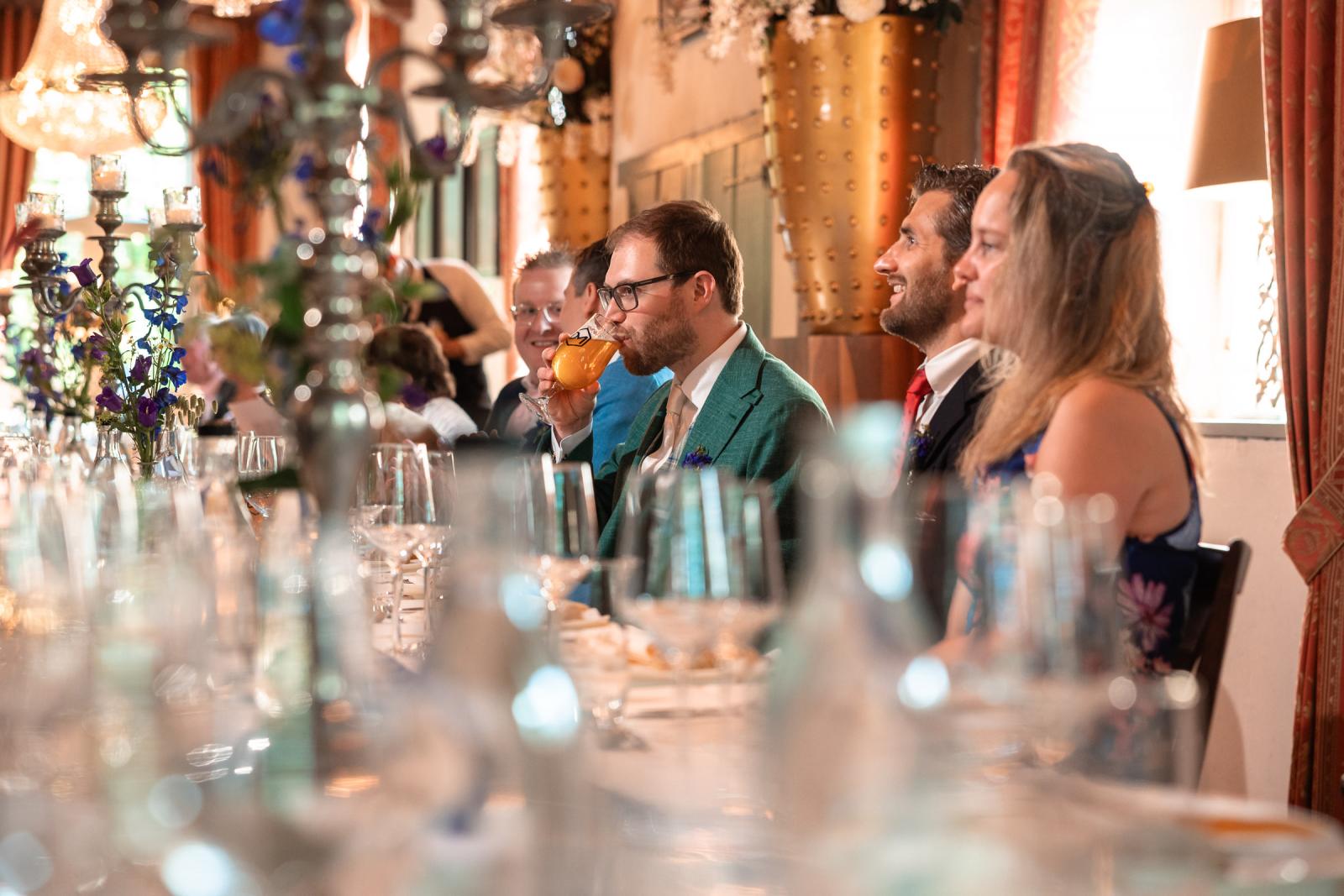 Bruidegom neemt een slok van zijn speciaalbier tijdens het trouwdiner, gefotografeerd met een sfeervolle, onscherpe voorgrond van glazen