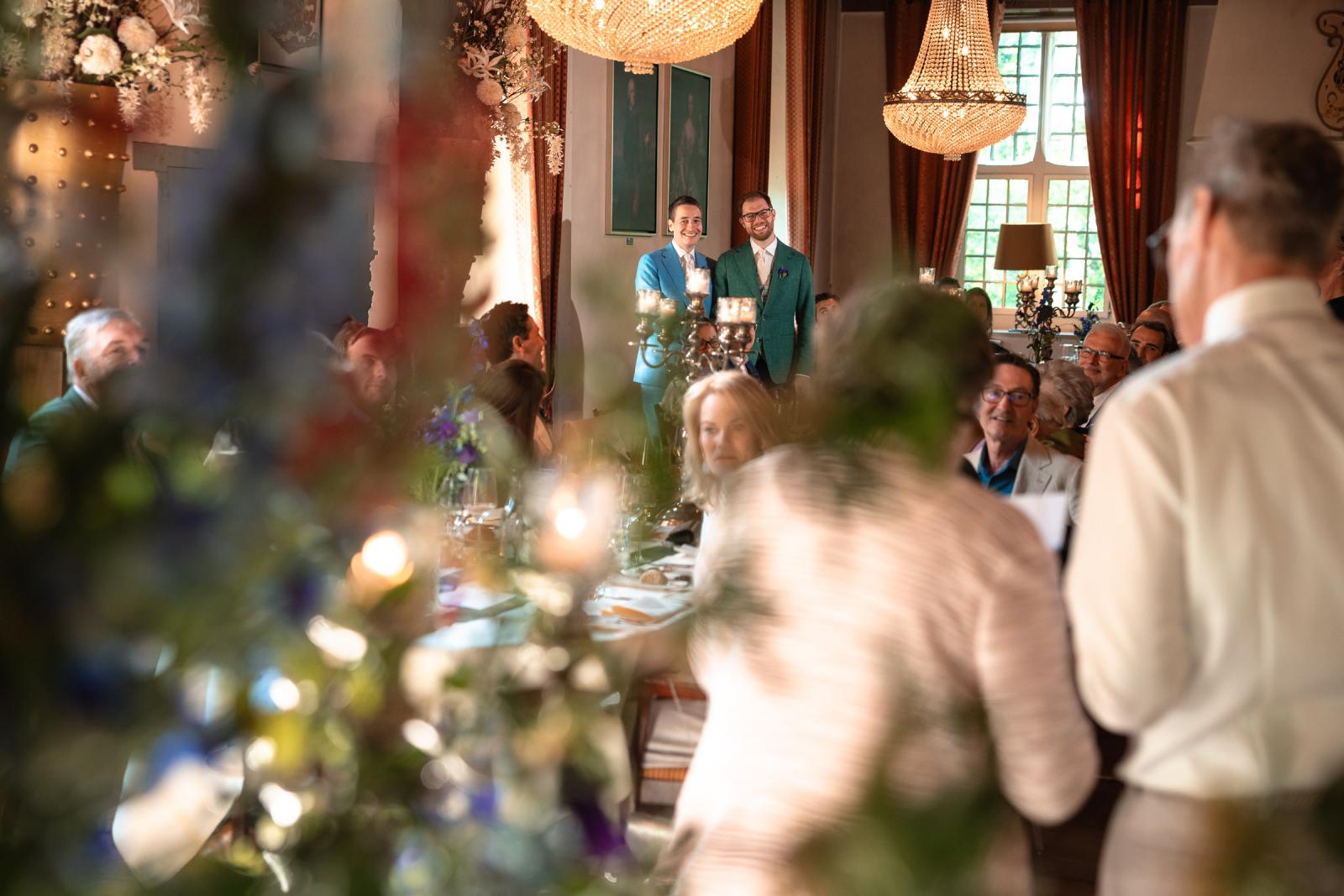 Daan en Martijn staan glimlachend achterin de ridderzaal terwijl een van de gasten, gezien op de rug, een speech voor hen houdt tijdens het diner