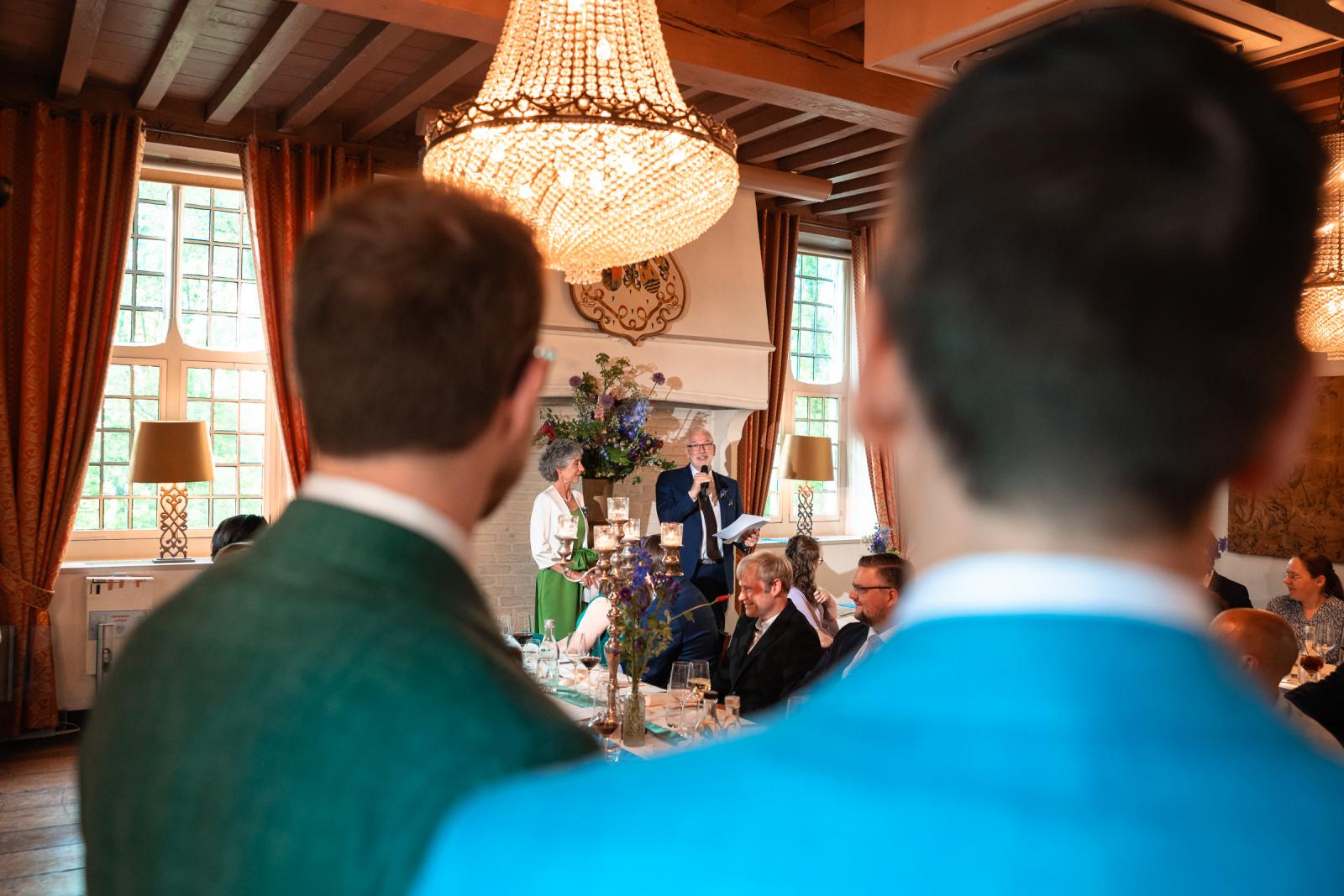 Blik over de schouders van het bruidspaar naar een oudere man die met een microfoon een speech houdt aan de dinertafel onder een grote kristallen kroonluchter