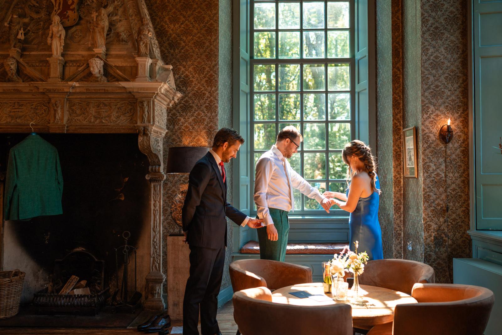 Martijn past zijn trouwpak in de salon van het kasteel, terwijl twee daggasten hem assisteren bij de mouwen van zijn overhemd