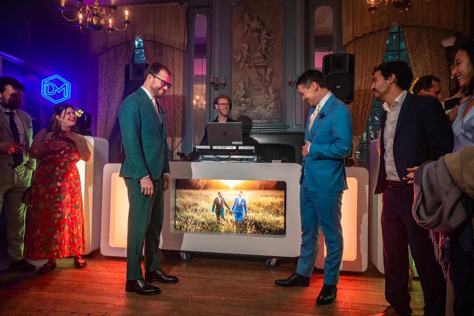 Daan en Martijn staan lachend tegenover elkaar bij de DJ-booth, waarop een verlicht scherm een foto van hun fotosessie in het grasveld toont