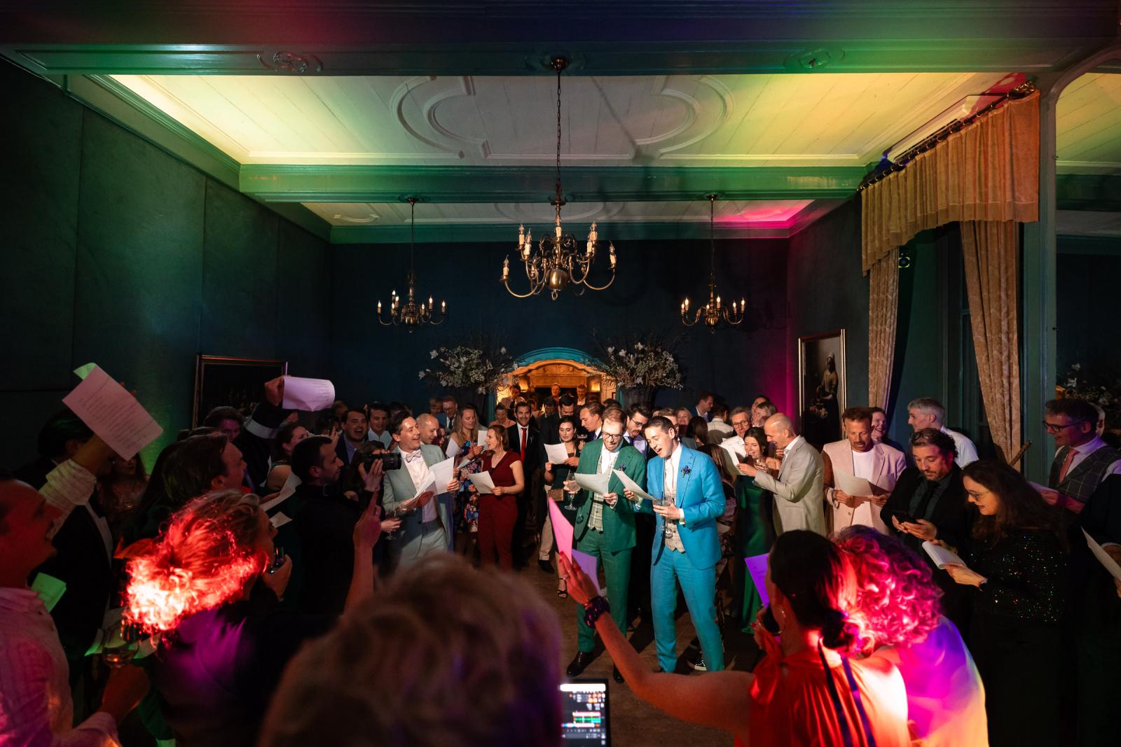 Overzichtsbeeld van de goedgevulde, sfeervol verlichte zaal waarin het bruidspaar en de gasten met papieren in de hand samen een lied zingen