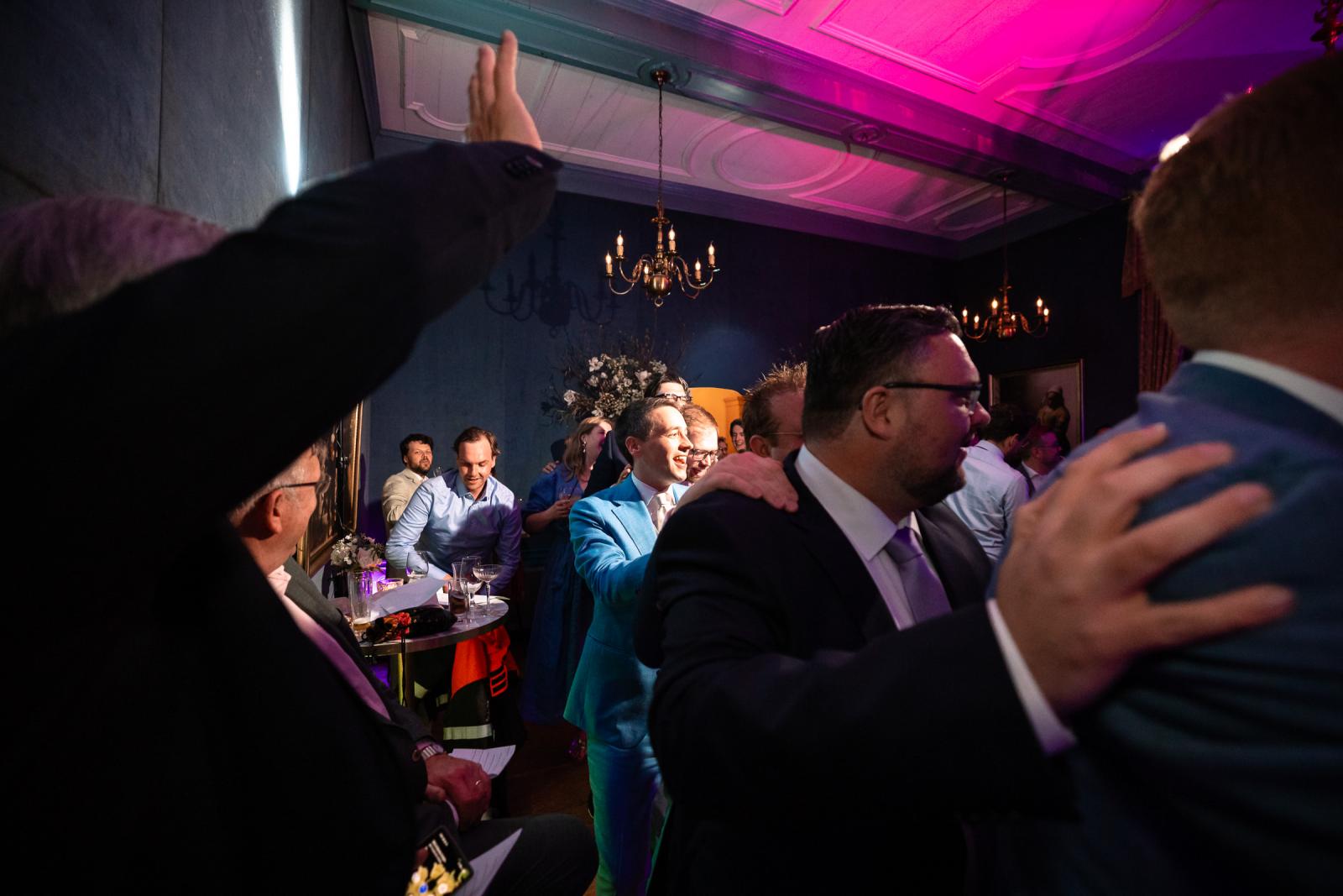 Daan loopt lachend in zijn blauwe pak door de feestende menigte terwijl een gast zijn hand opsteekt en anderen hem op de schouders kloppen