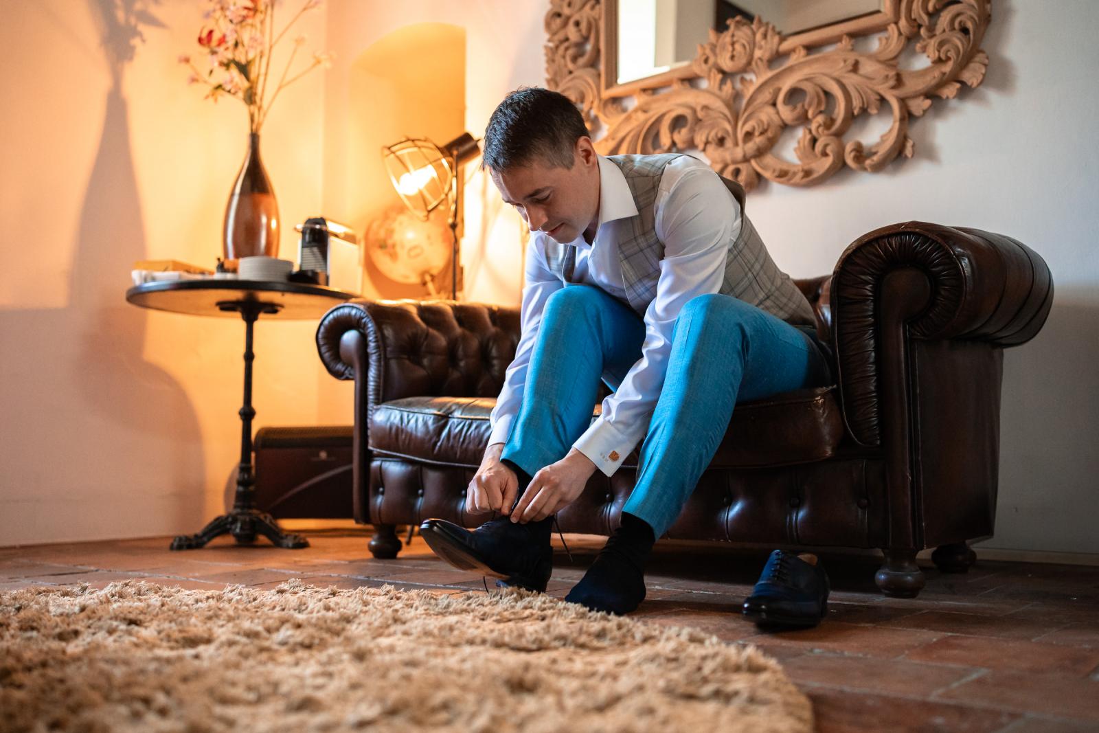 Bruidegom Daan zit op een gecapitonneerde leren Chesterfieldbank en strikt de veters van zijn donkerblauwe trouwschoenen
