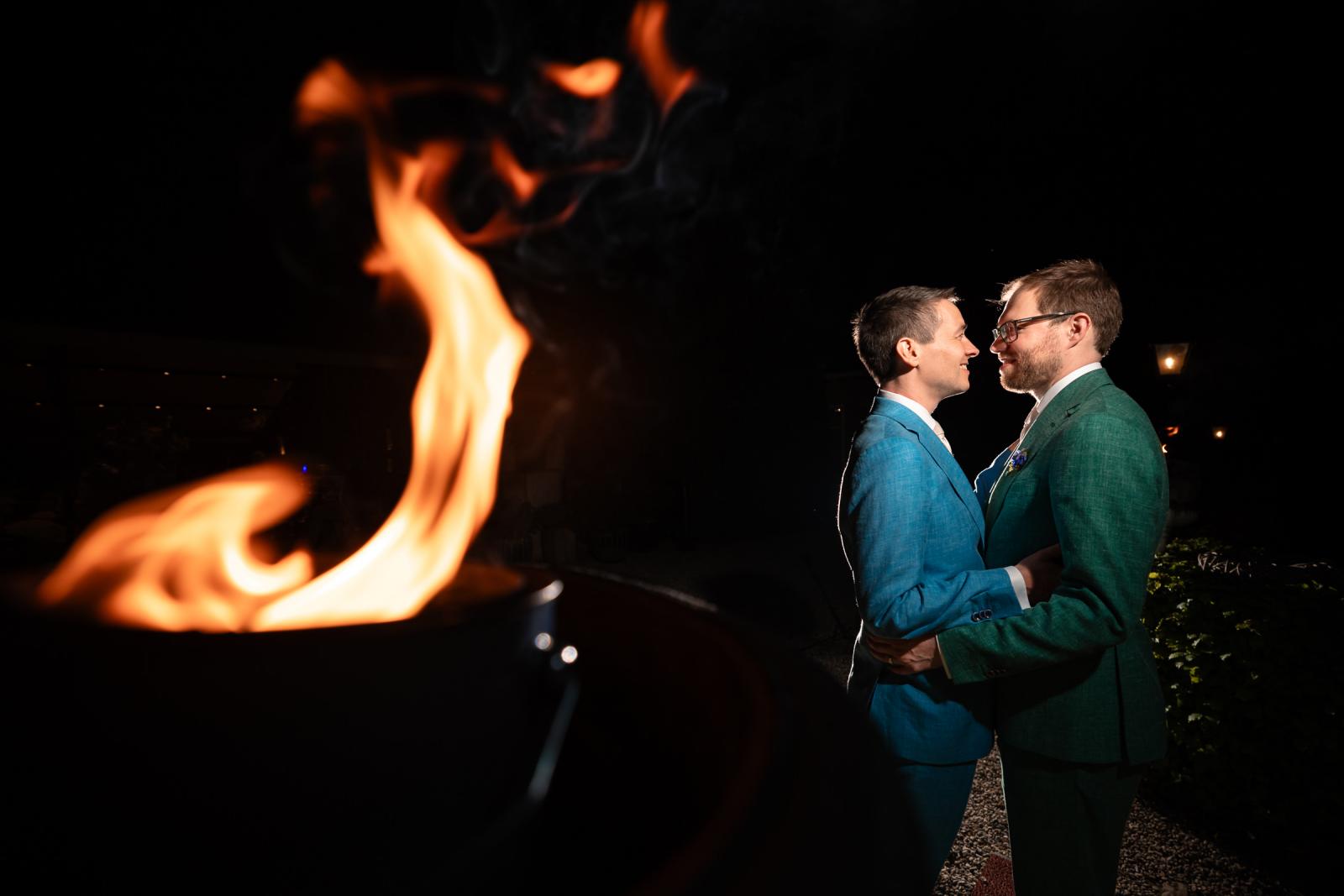 Close-up avondportret van Daan en Martijn die elkaar teder aankijken, met op de voorgrond de sfeervolle, onscherpe vlammen van een vuurkorf