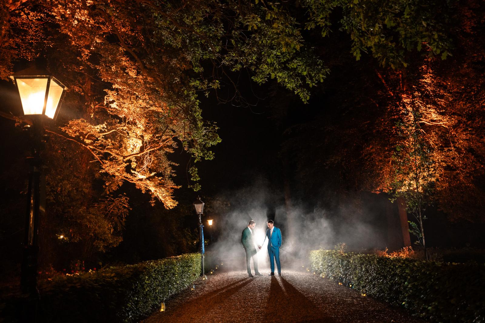Sfeervol avondportret van Daan en Martijn die hand in hand op een grindpad staan, omringd door een mysterieuze rookwolk en dramatisch tegenlicht dat lange schaduwen naar voren werpt onder de bomen