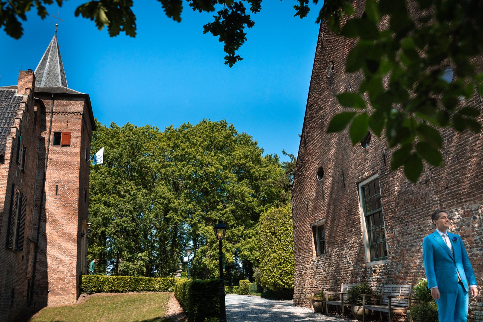 Bruidegom Dan staat in zijn blauwe trouwpak buiten te wachten bij de kasteelmuur voor het 'first look'-moment met Martijn