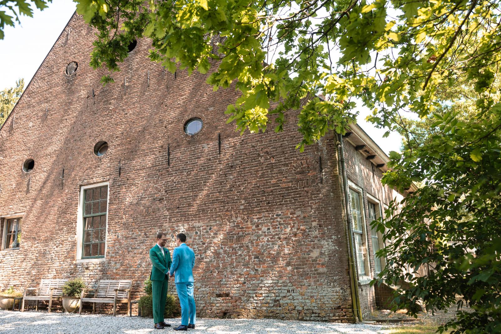 Martijn in zijn groene pak en Daan in zijn blauwe pak hebben hun 'first look' en ontmoeten elkaar glimlachend buiten bij Kasteel Wijenburg