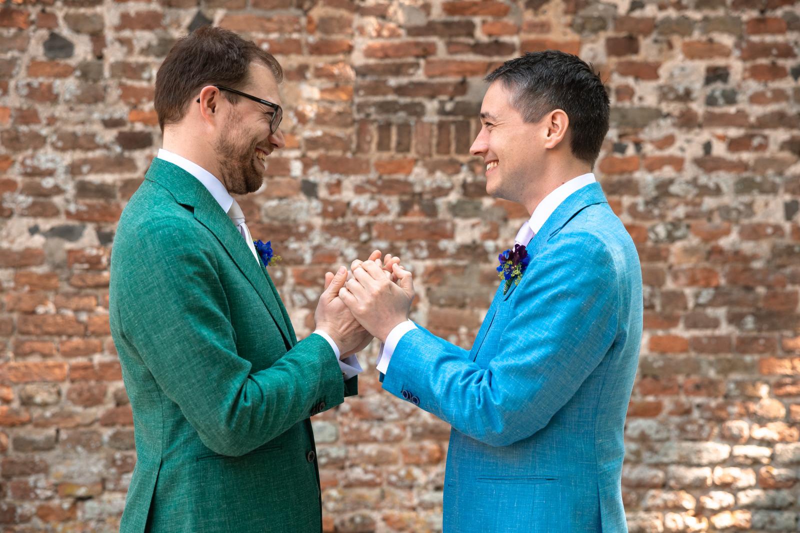 Close-up van Daan en Martijn die elkaars handen vasthouden en breed naar elkaar lachen met een oude bakstenen kasteelmuur op de achtergrond