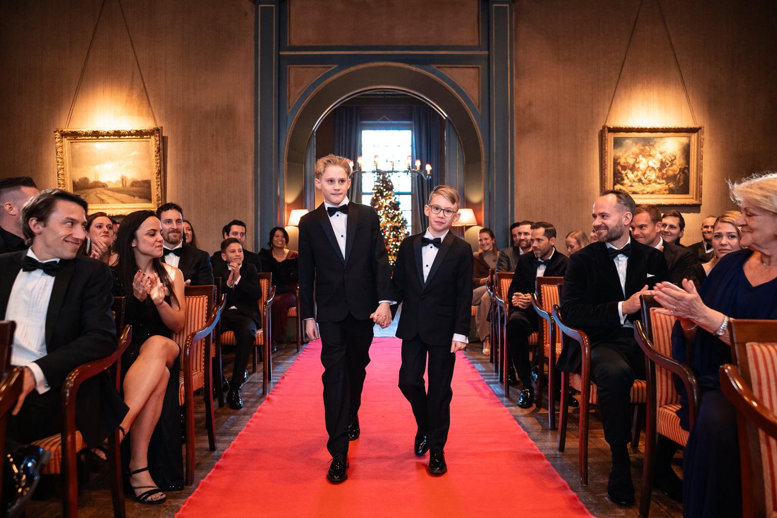 De zoons lopen hand in hand de ceremonie binnen, kerstversiering op de achtergrond.