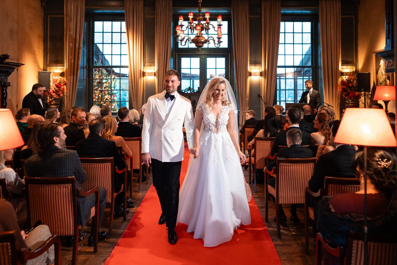 Bruidspaar loopt hand in hand weg na de ceremonie, gasten applaudisseren.