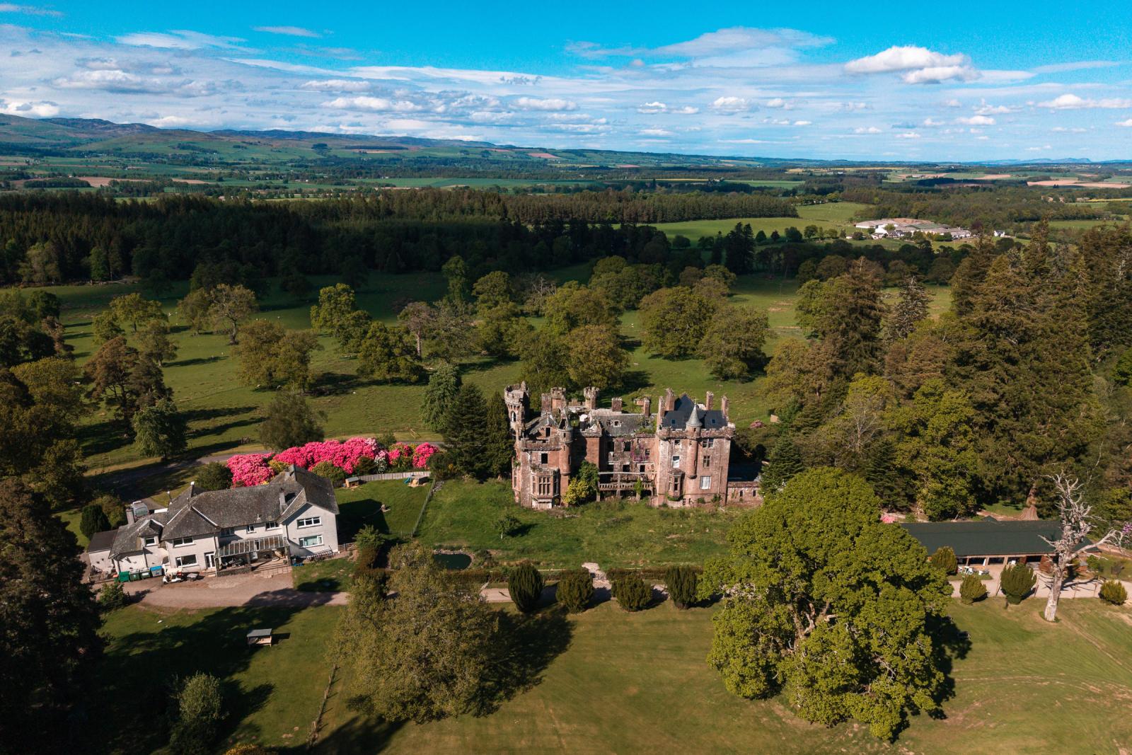 Aerial drone view of Culdees Castle Estate wedding venue in Perthshire Scotland, showcasing the historic stone castle ruins surrounded by lush green forests and vibrant blooming spring gardens.