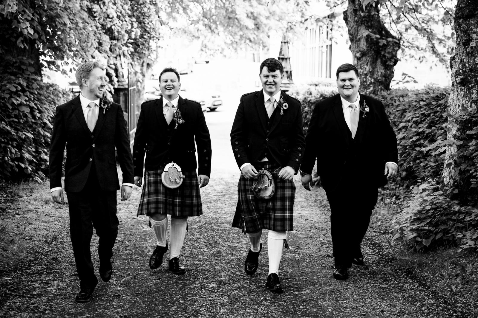 Black and white photograph of groom Hamish and his groomsmen in traditional Scottish attire laughing and walking down a tree-lined path towards Muthill church.