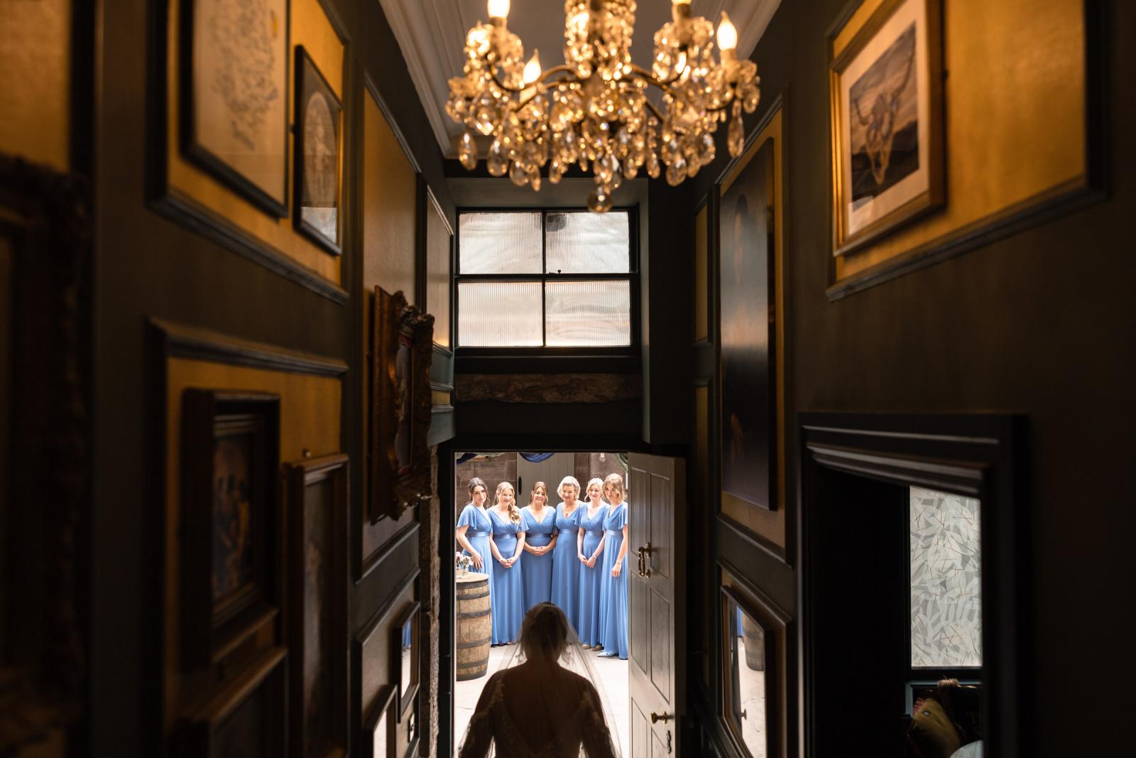 Interior perspective looking down a moody castle hallway lit by a crystal chandelier, with bride Emma walking toward her bridesmaids waiting outside the door.