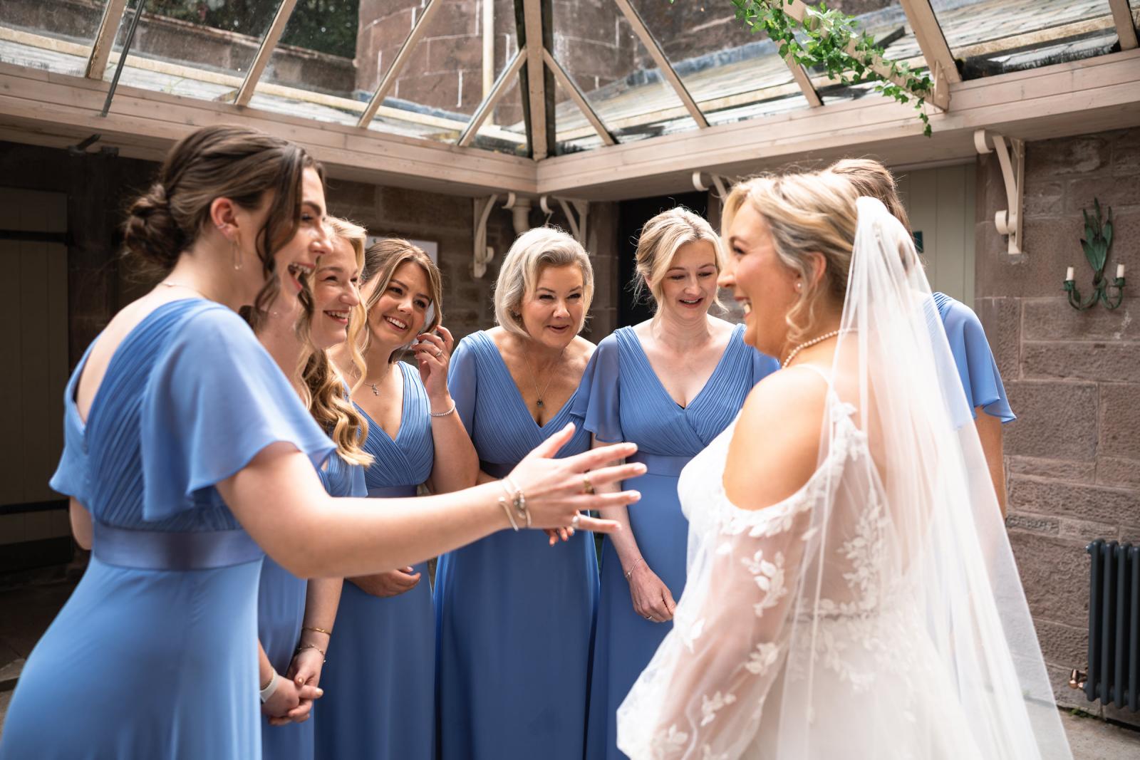 Candid moment of bridesmaids in cornflower blue dresses laughing and crying during a first look with bride Emma under a glass conservatory roof.