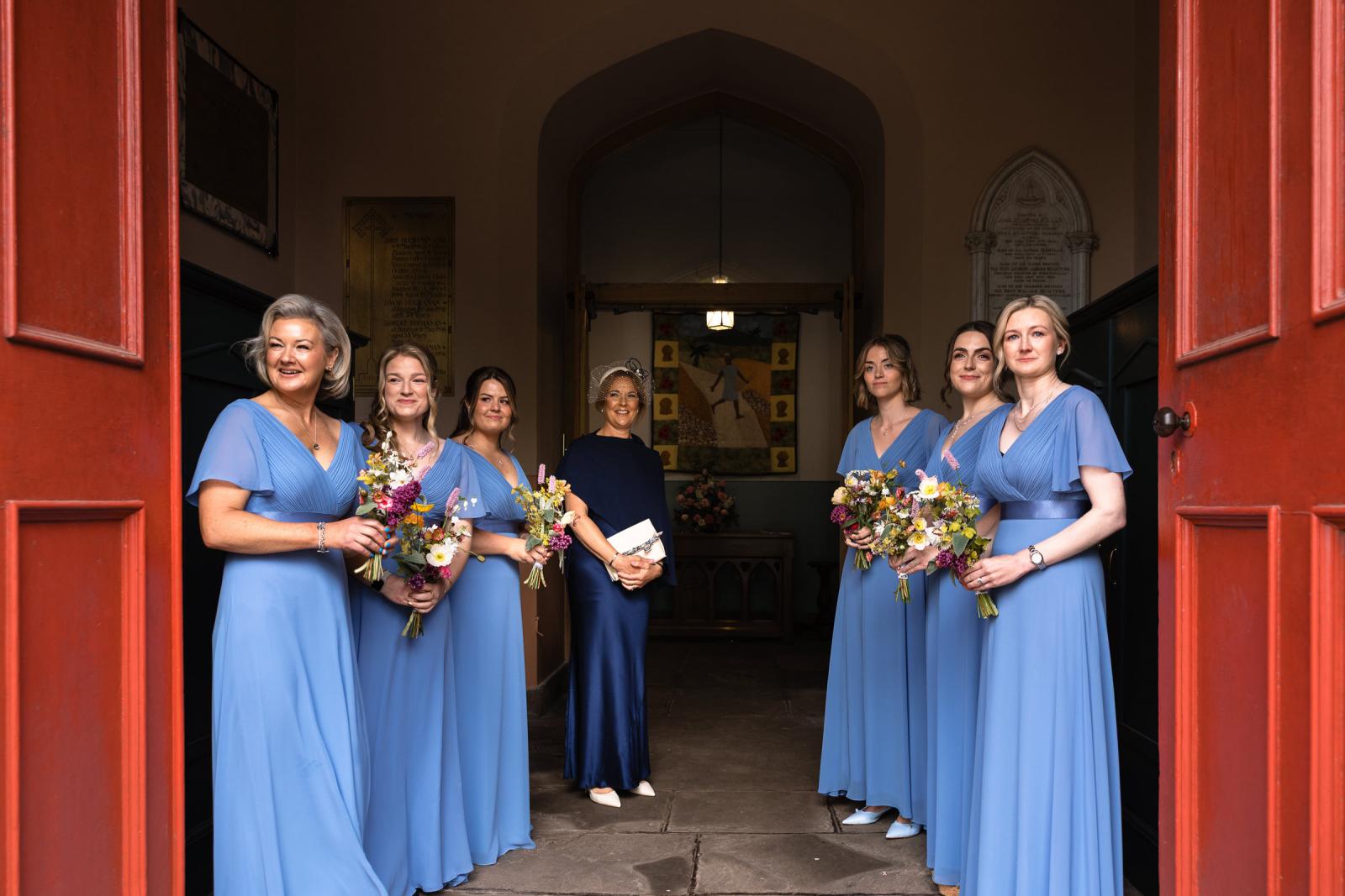 Bridesmaids in flowing blue gowns holding seasonal flowers while waiting inside the red double doors of the historic Muthill church.