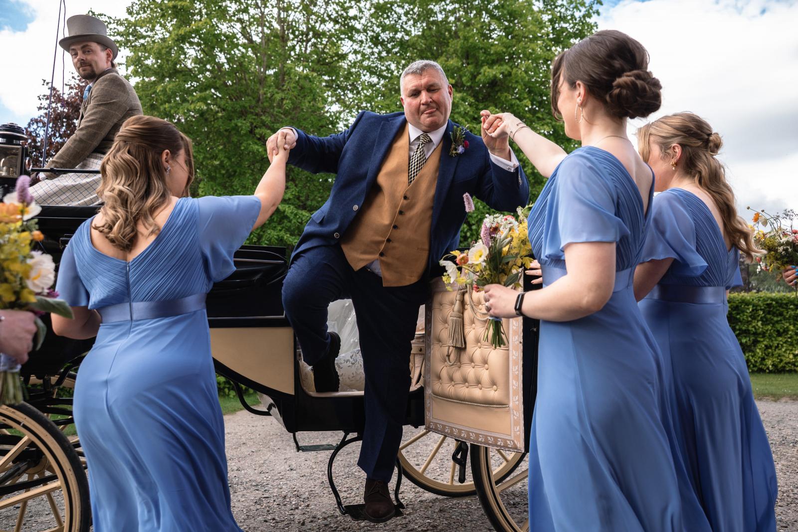 Emma’s father wearing a blue suit and gold waistcoat stepping out of a vintage horse-drawn wedding carriage with the help of the bridesmaids.
