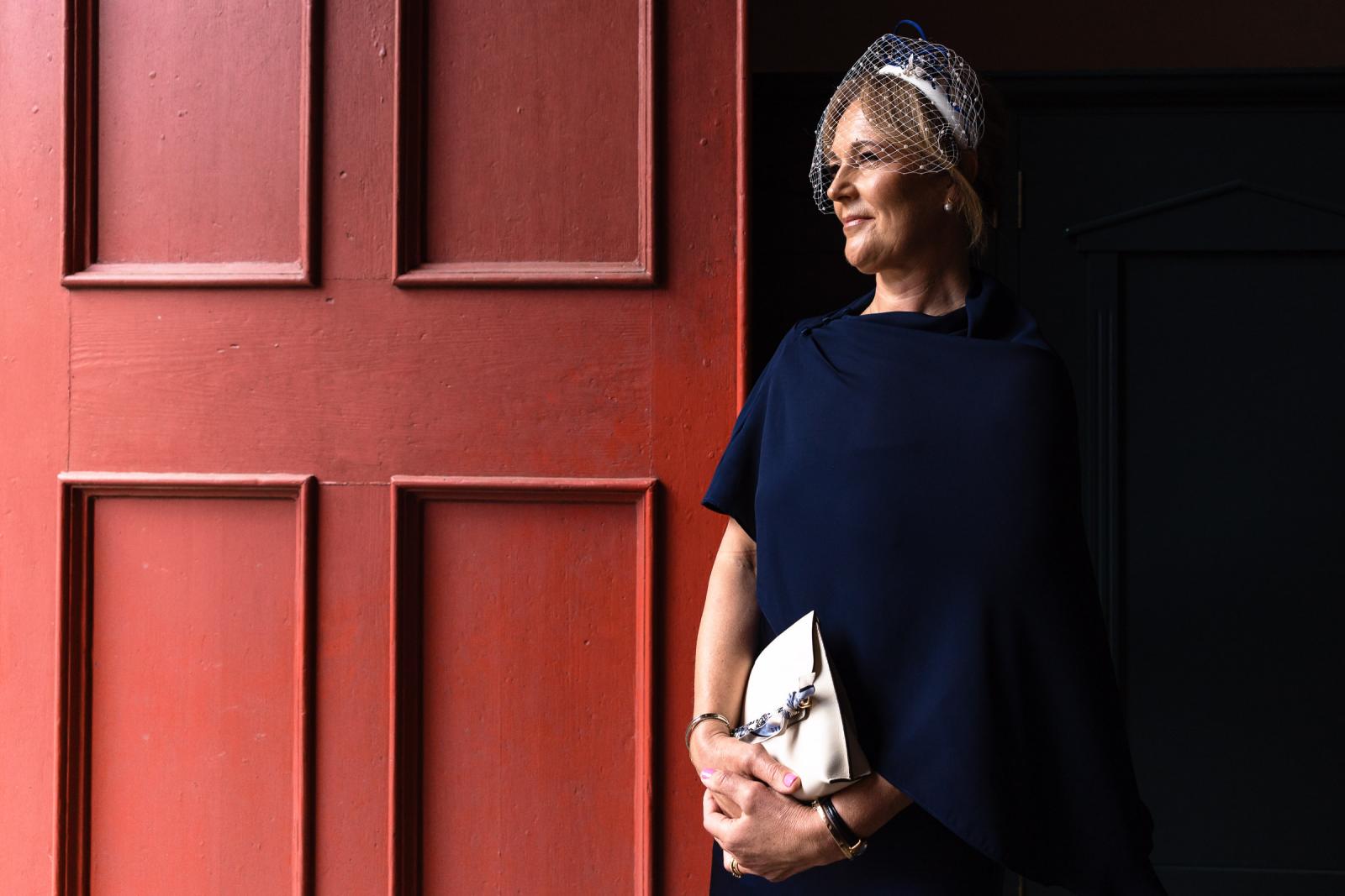 Elegant portrait of the mother of the bride dressed in a navy blue dress and a matching birdcage veil, standing gracefully by a large red wooden church door.