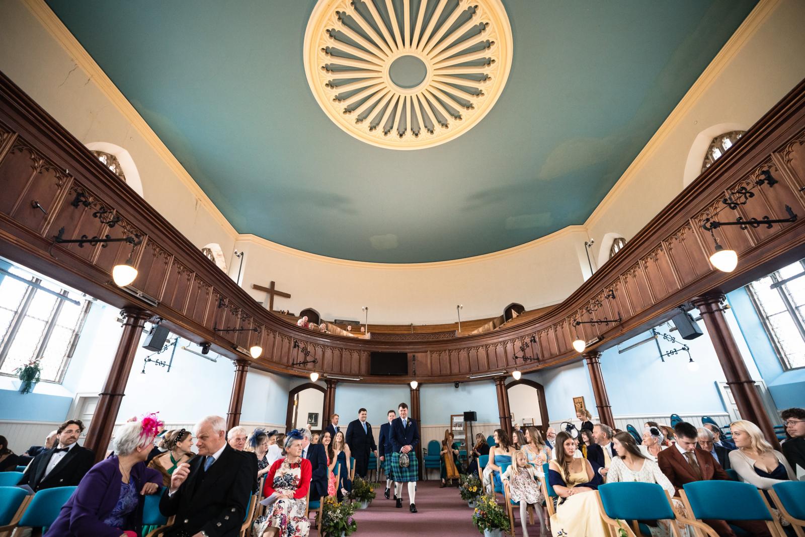 Wide interior shot of the circular Scottish church in Muthill, capturing groom Hamish walking down the aisle towards the front before the ceremony begins.