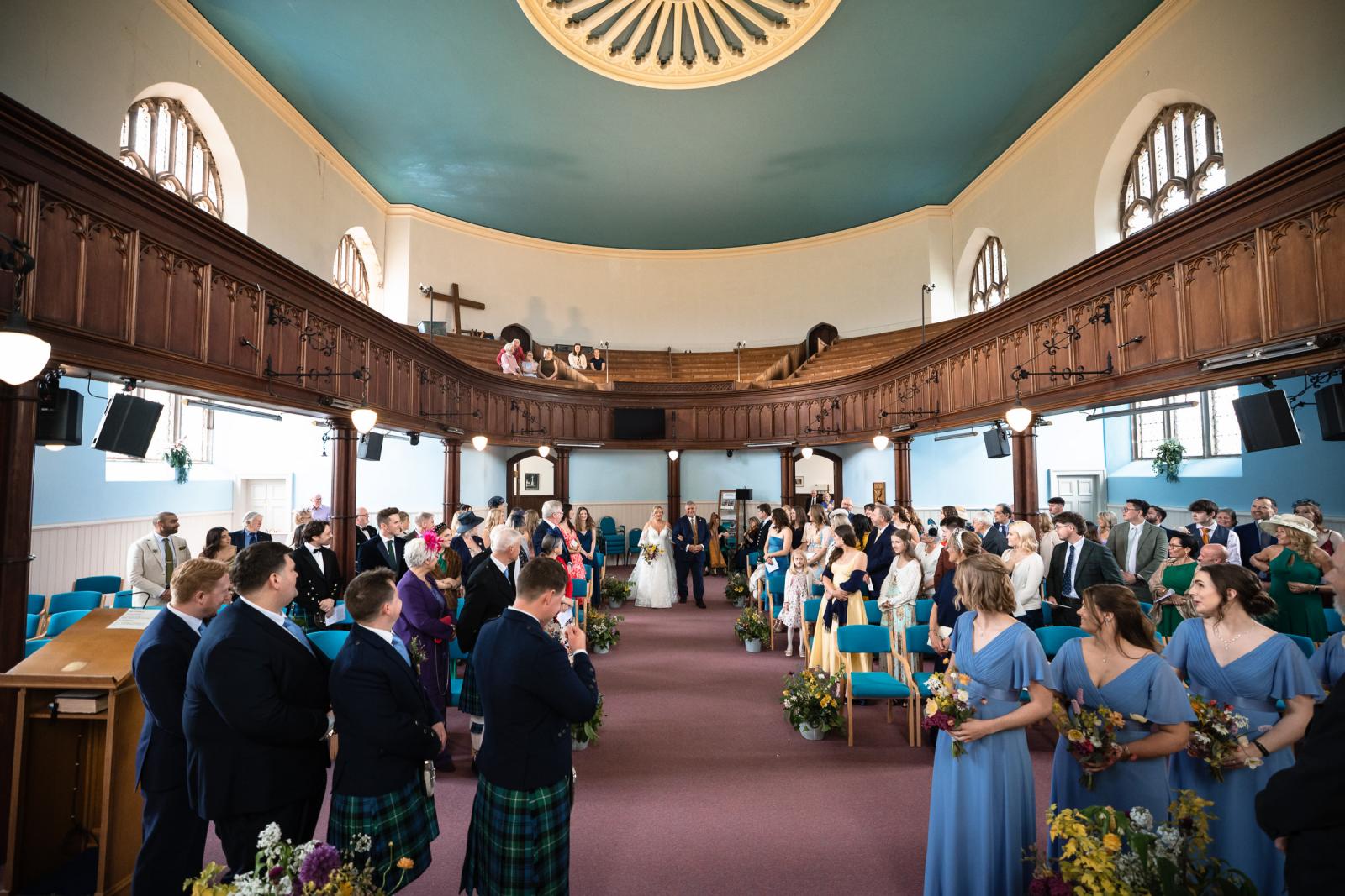 Wide-angle perspective of the indoor church wedding ceremony in Muthill, showing the bride walking down the aisle with her father as guests look on from the pews and balcony.