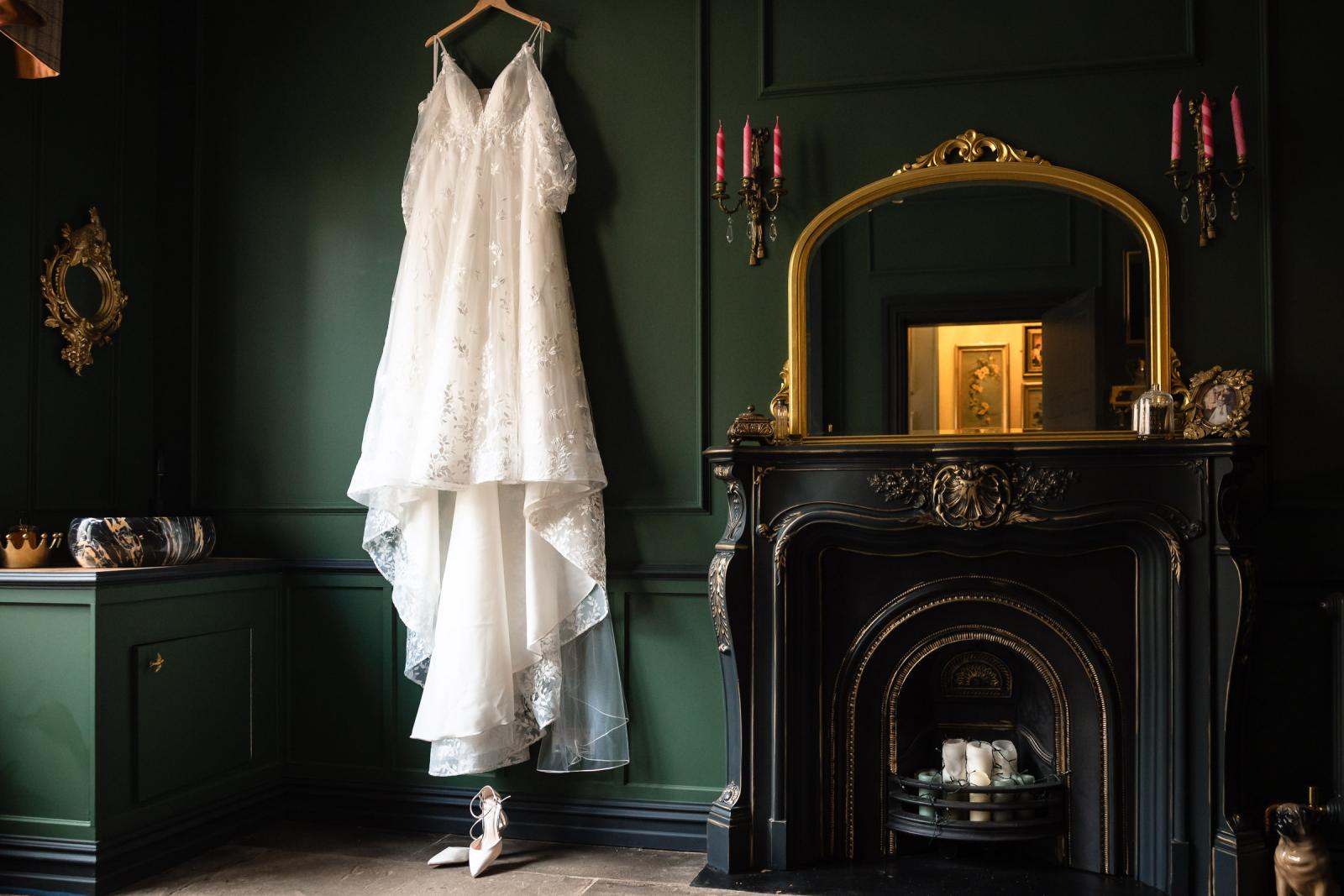 Emma’s off-the-shoulder lace wedding dress hanging on a dark green wall next to an ornate gold mirror and vintage black fireplace mantle at Culdees Castle Estate.