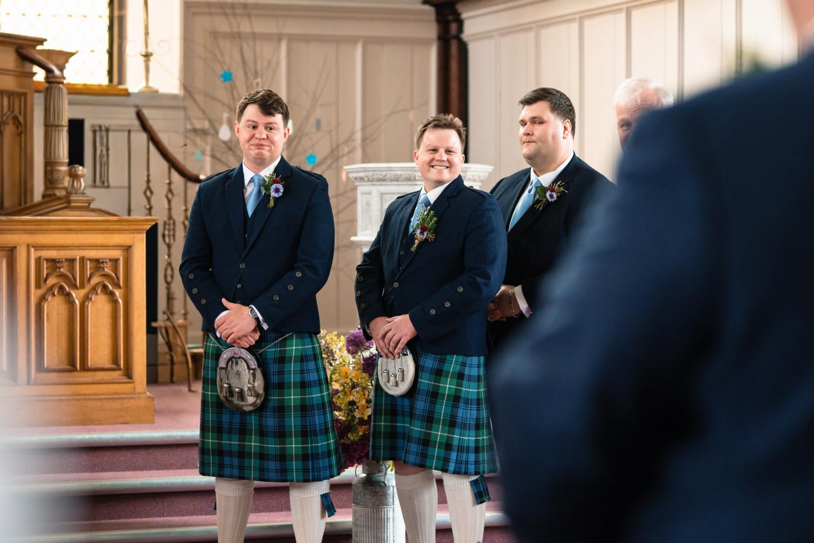 Close-up of a groom in a traditional Scottish kilt standing proudly at the altar, smiling warmly with tears in his eyes alongside his best man and groomsman during the wedding ceremony.