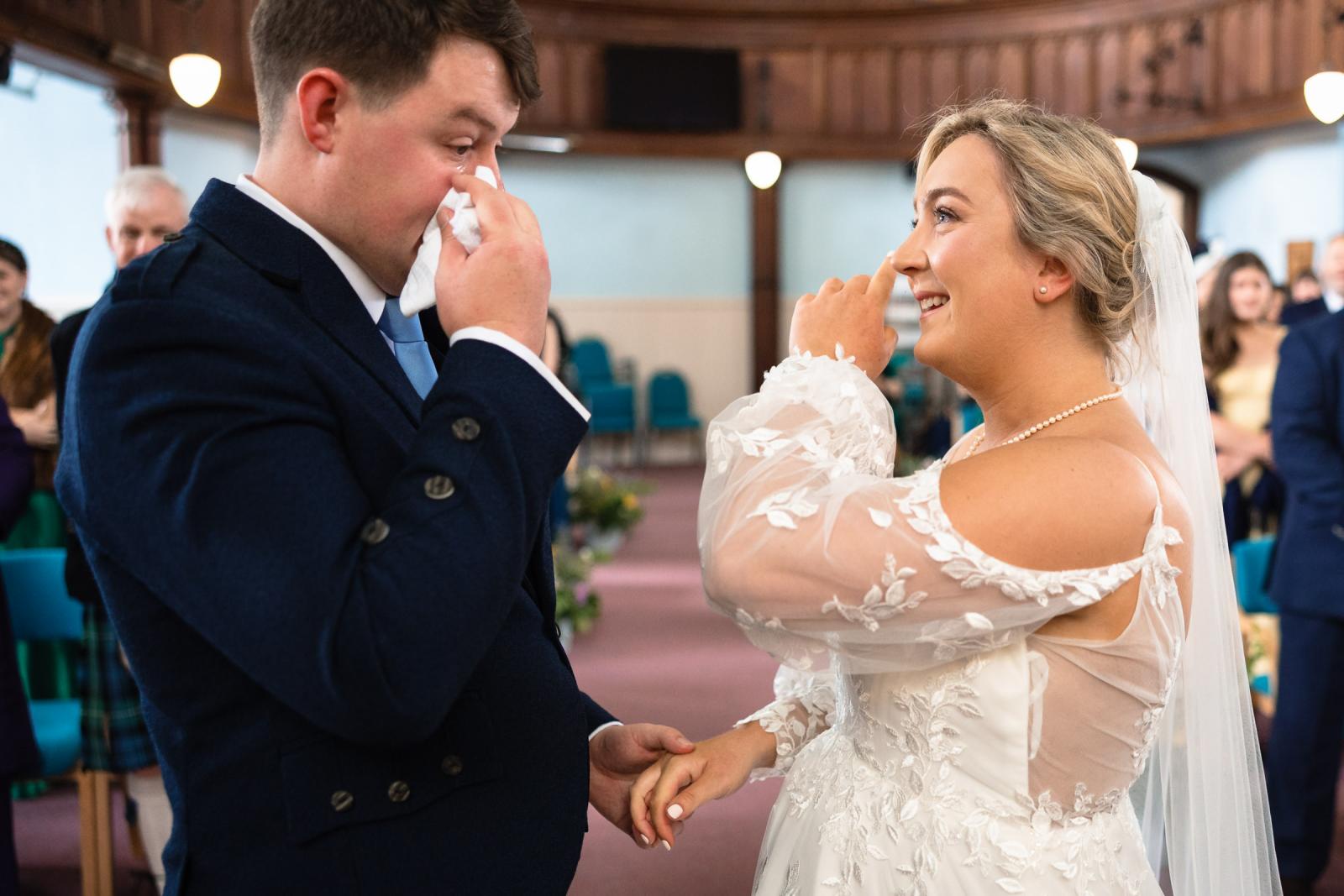Close-up emotional portrait of groom Hamish wiping a tear with a handkerchief while bride Emma smiles lovingly and wipes away her own tear during their Muthill church wedding ceremony.
