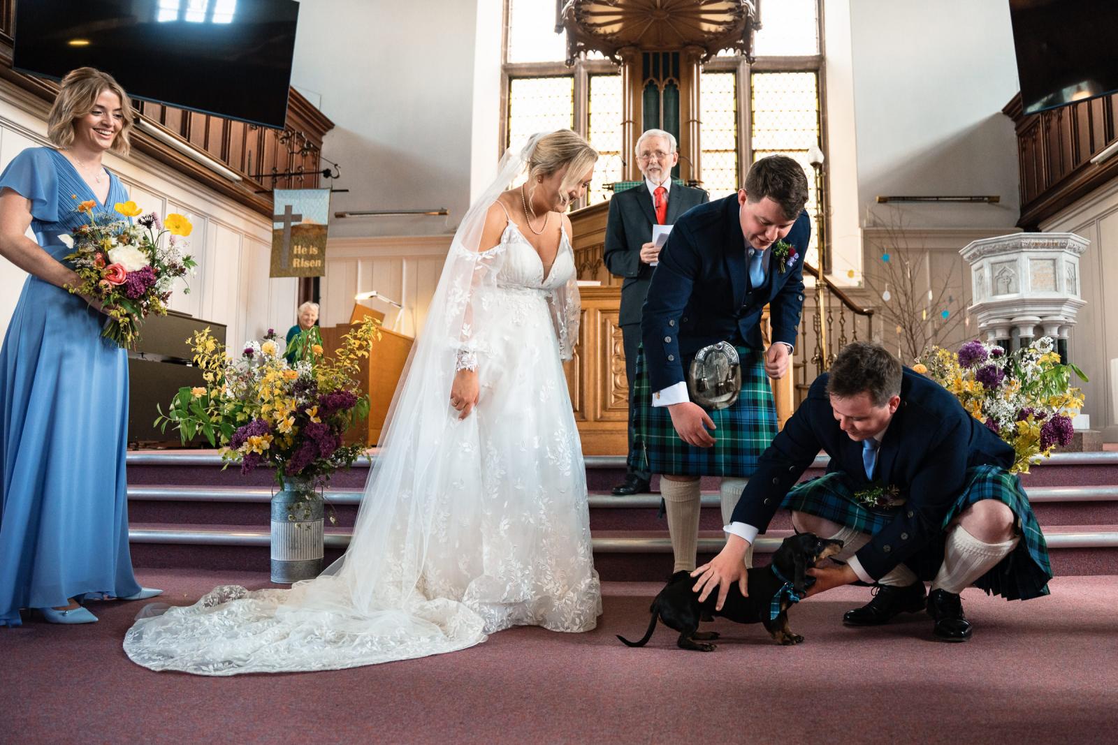 The groom’s brother kneels down at the altar to catch the dachshund ring bearer as bride Emma and groom Hamish laugh during their playful church wedding service.