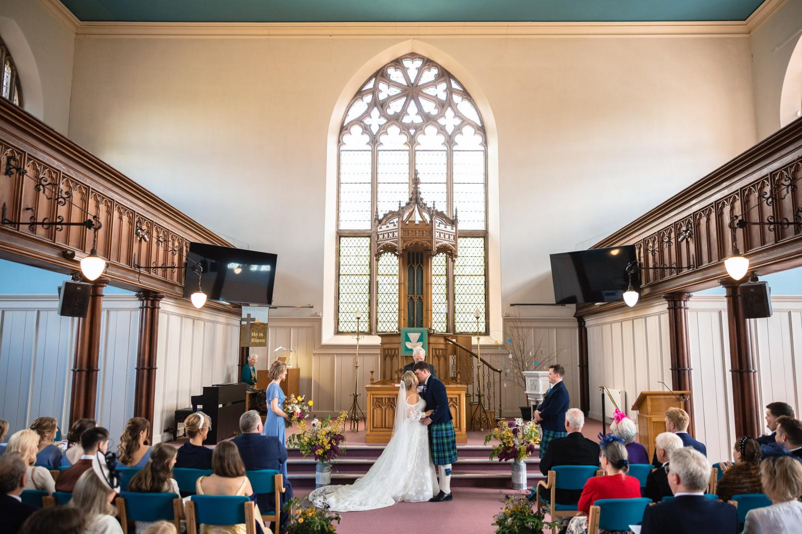 Wide-angle interior photograph of Muthill church showing Emma and Hamish sharing their first kiss at the altar, surrounded by guests in pews and grand wooden balconies.
