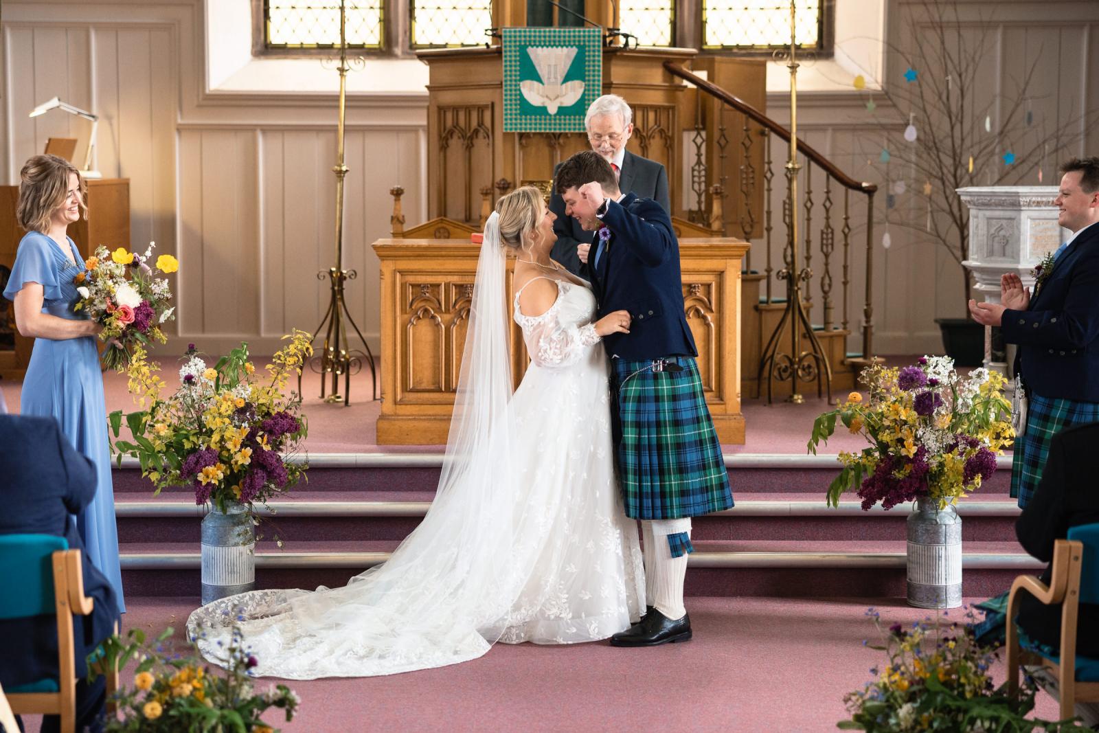 Groom Hamish victoriously throws his hand in the air with pure joy right after their first kiss as husband and wife, standing next to a smiling Emma at the altar.
