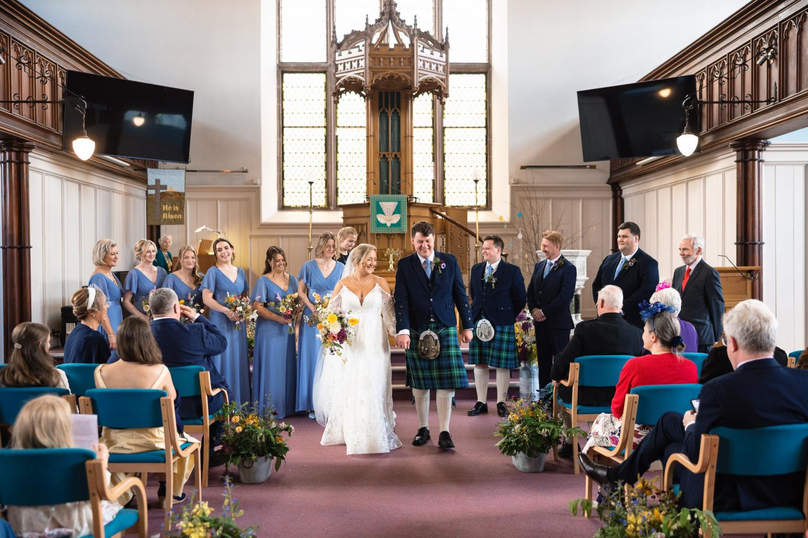 The newlyweds Emma and Hamish walk down the church aisle hand-in-hand as husband and wife, followed by their bridesmaids in blue dresses and groomsmen in kilts.