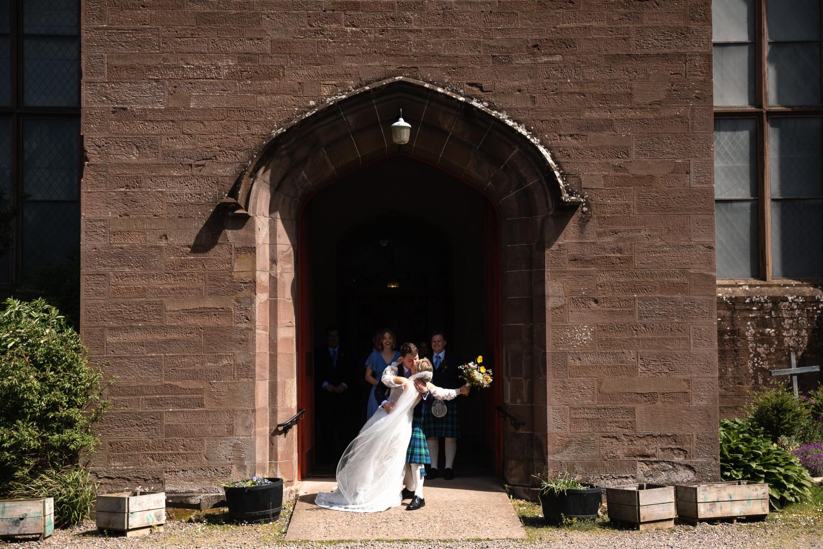 Groom Hamish dips bride Emma for a romantic kiss underneath the large stone archway of the Muthill church doors as guests watch from behind.