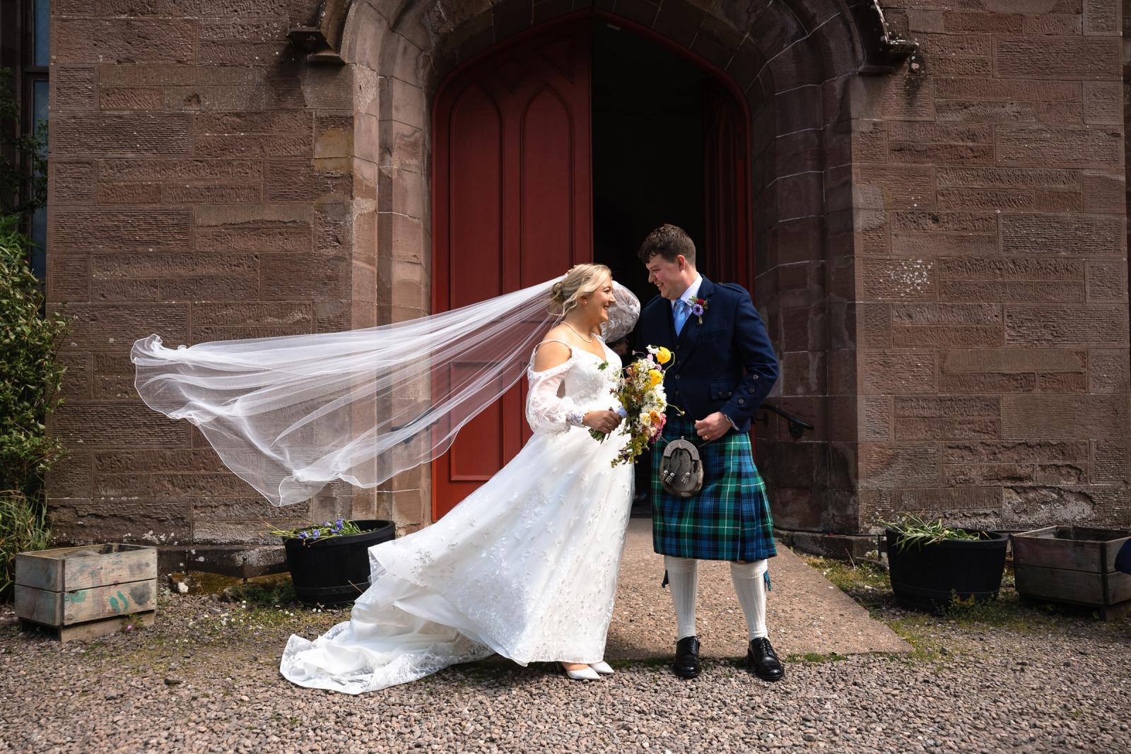 Emma and Hamish smiling and standing outside the red church doors in Perthshire, Scotland as a sudden gust of wind dramatically blows the bride’s long tulle wedding veil.