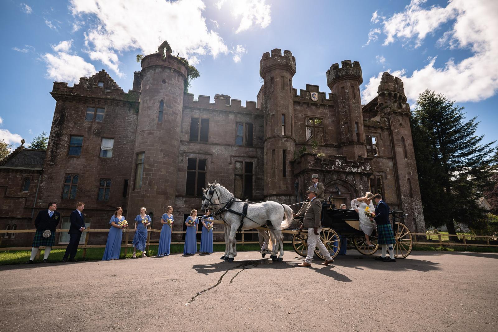 Emma and Hamish step out of a luxury white horse-drawn carriage in front of the historic, sunlit stone ruins of Culdees Castle Estate as bridesmaids and groomsmen line up to welcome them.