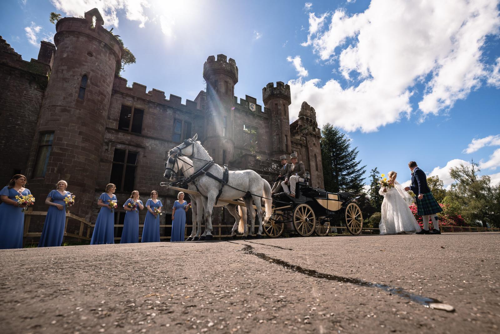 Wide-angle shot of Emma and Hamish standing by their horse-drawn carriage under a blue sky, with bridesmaids lining up outside the majestic stone ruins of Culdees Castle Estate.