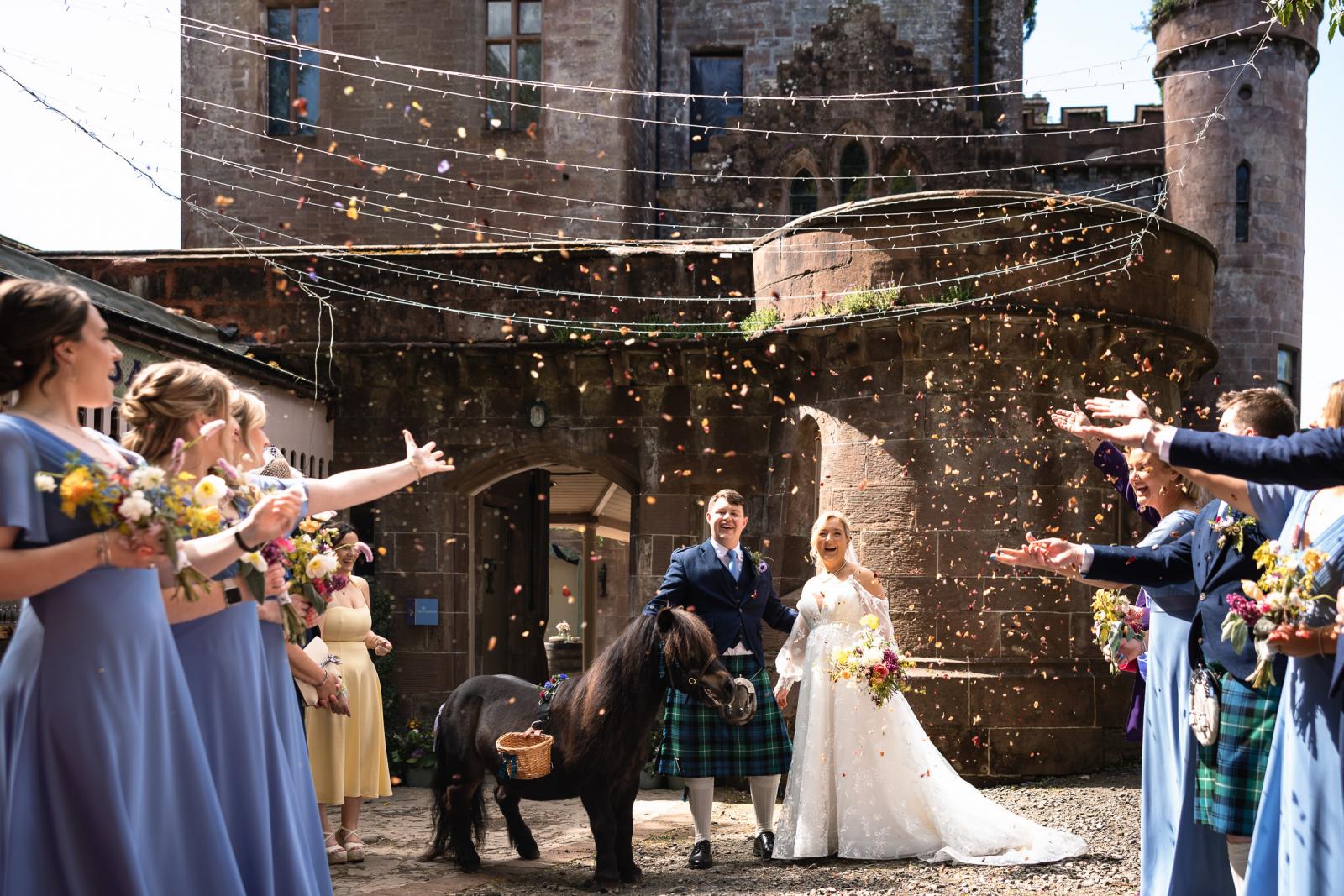 Emma and Hamish walking through a vibrant shower of dried flower confetti at Culdees Castle, accompanied by an adorable miniature pony wearing a flower garland and a small wicker basket.