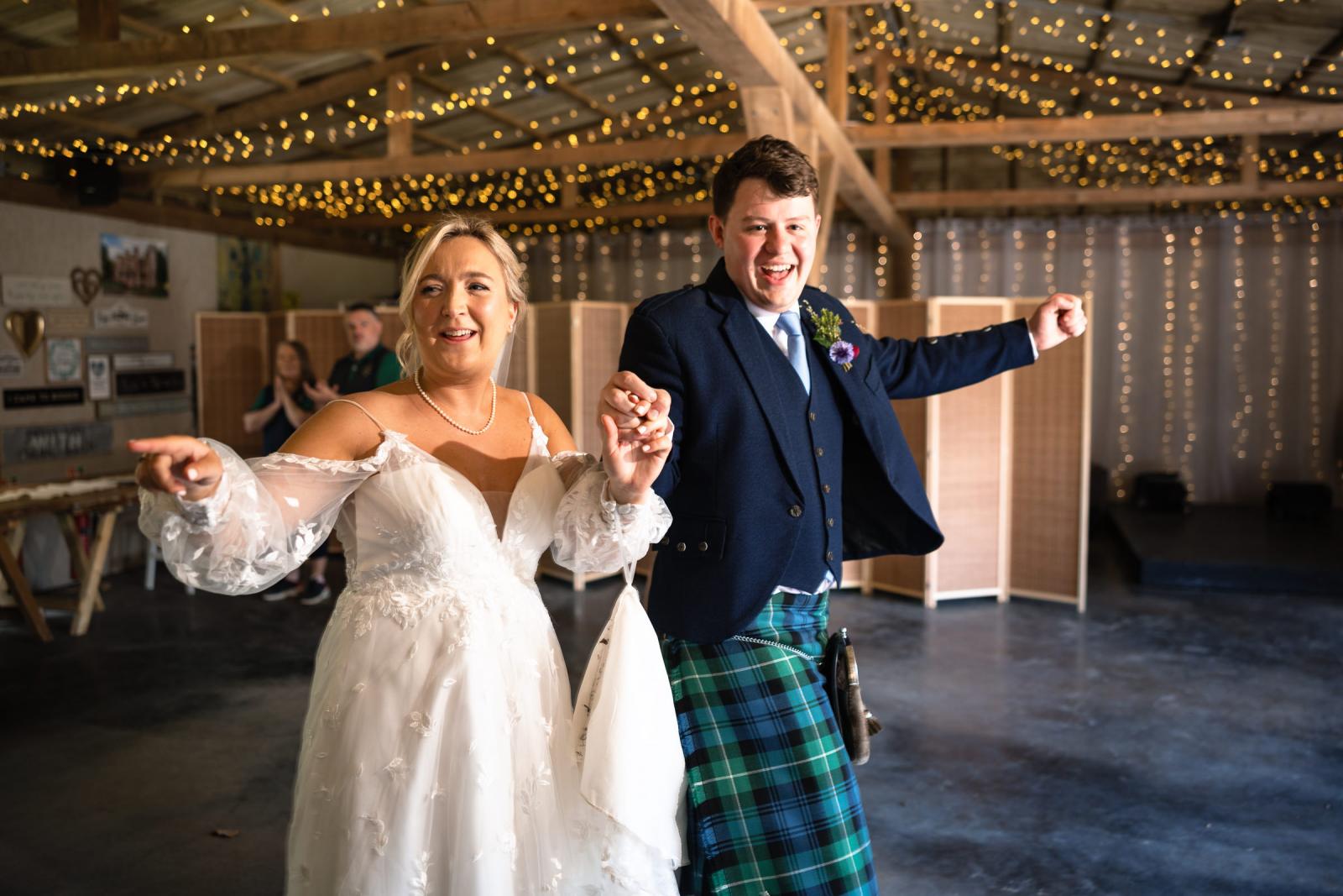 The newlyweds joyfully cheer and dance as they make their grand entrance into their wedding reception barn, beautifully decorated with thousands of twinkling fairy lights.