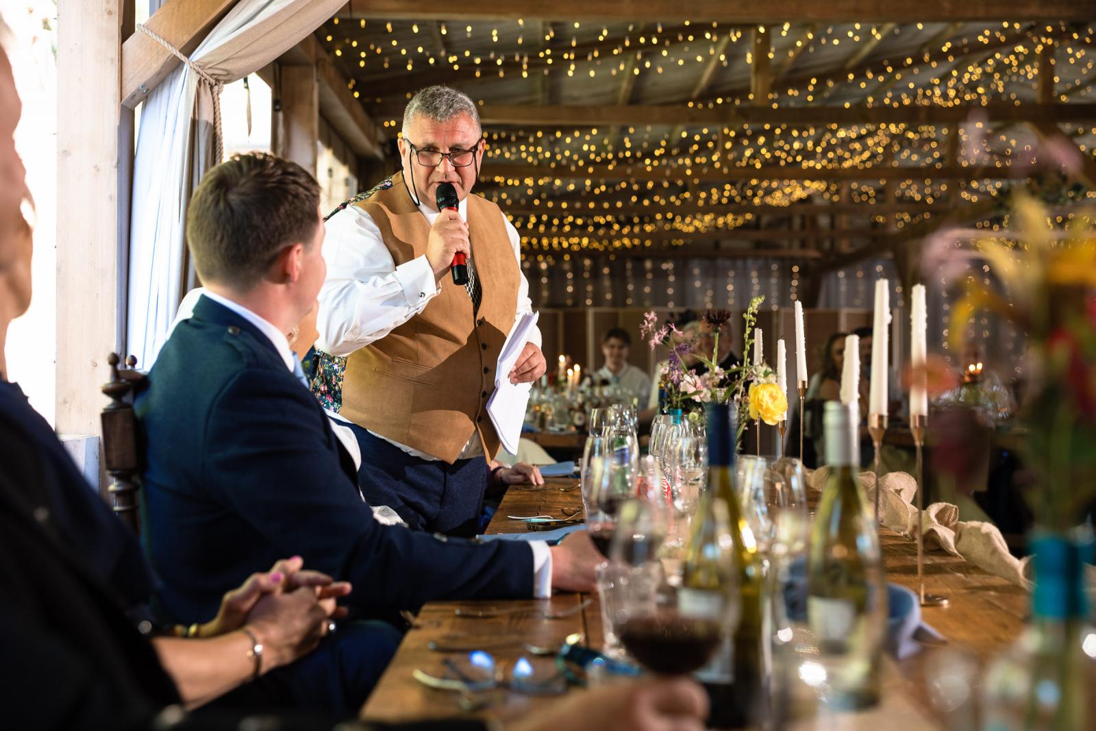 The father of the bride wearing a beige waistcoat stands up to deliver an emotional speech into a microphone at a rustic wooden top table adorned with candles and wild flowers.