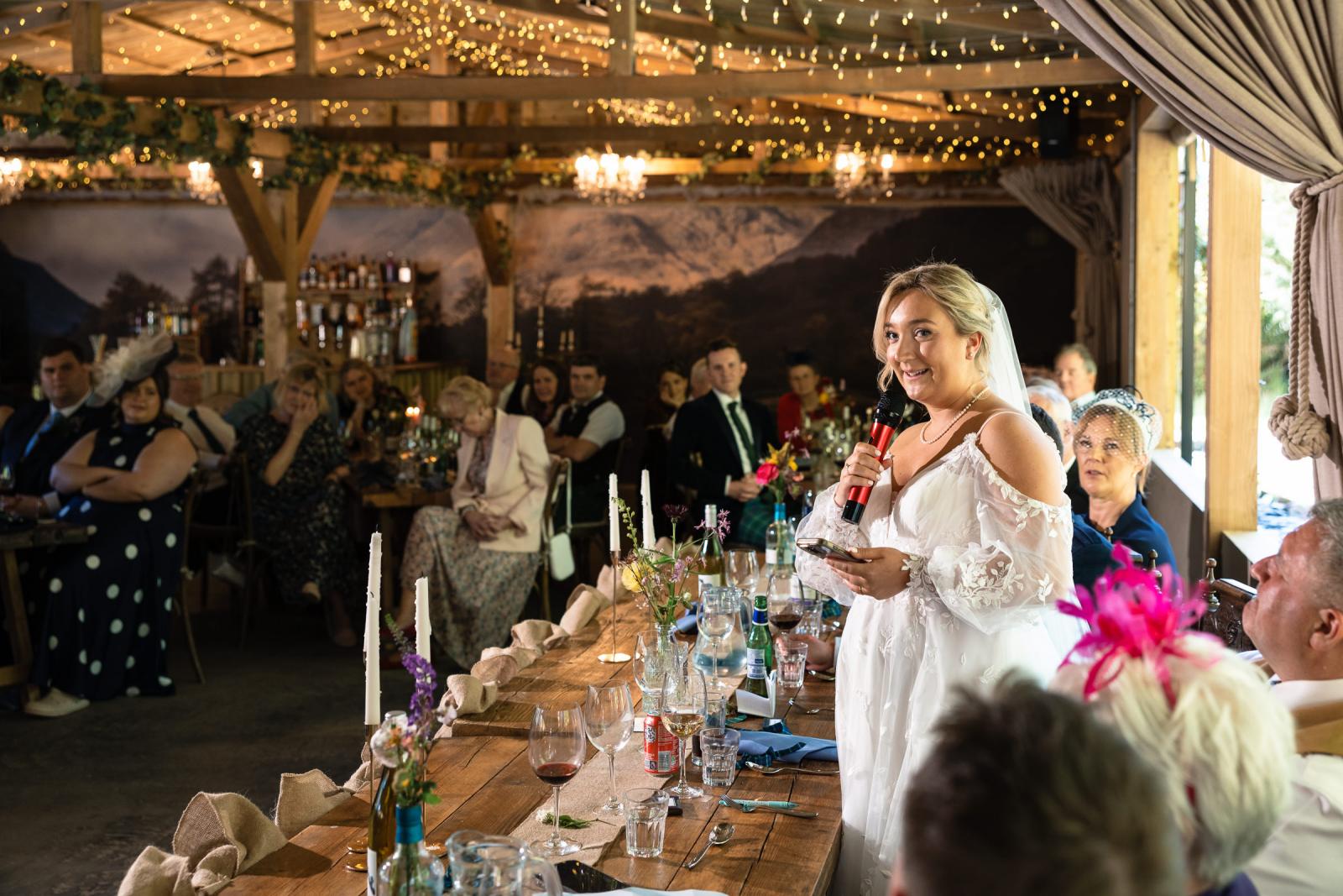 Bride Emma stands at the wedding top table, smiling warmly as she gives her bride speech into a microphone to a room full of seated wedding guests.