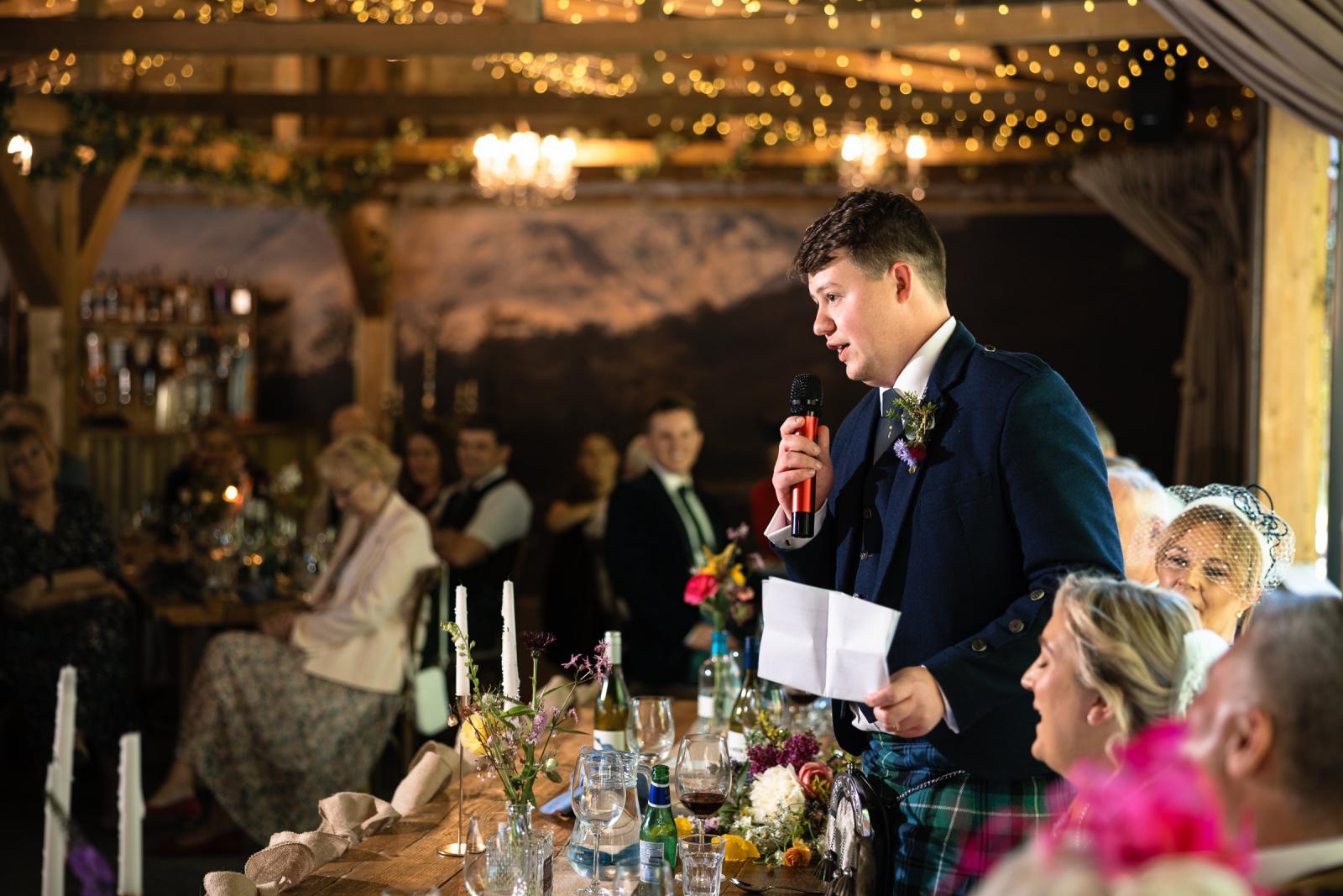Groom Hamish stands proudly at the rustic top table, reading from a piece of paper as he delivers his groom speech during the wedding dinner.