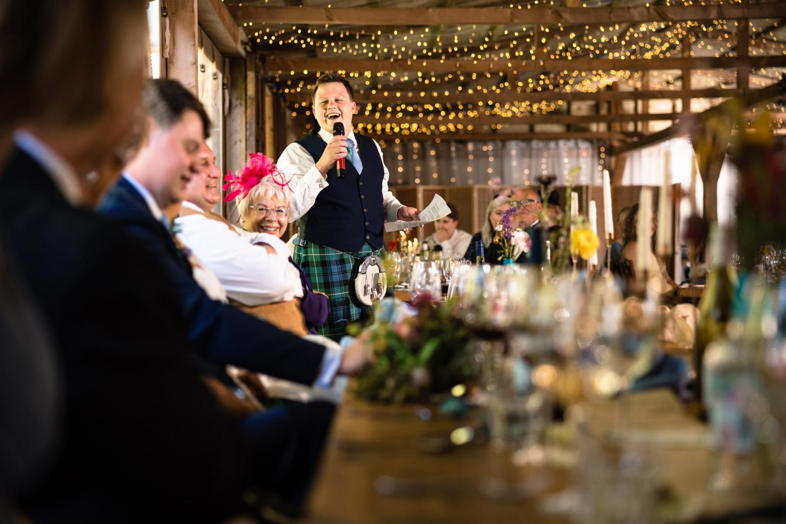 The best man in a traditional Scottish kilt stands up to deliver a hilarious speech, making the groom, bride, and wedding guests laugh heartily.